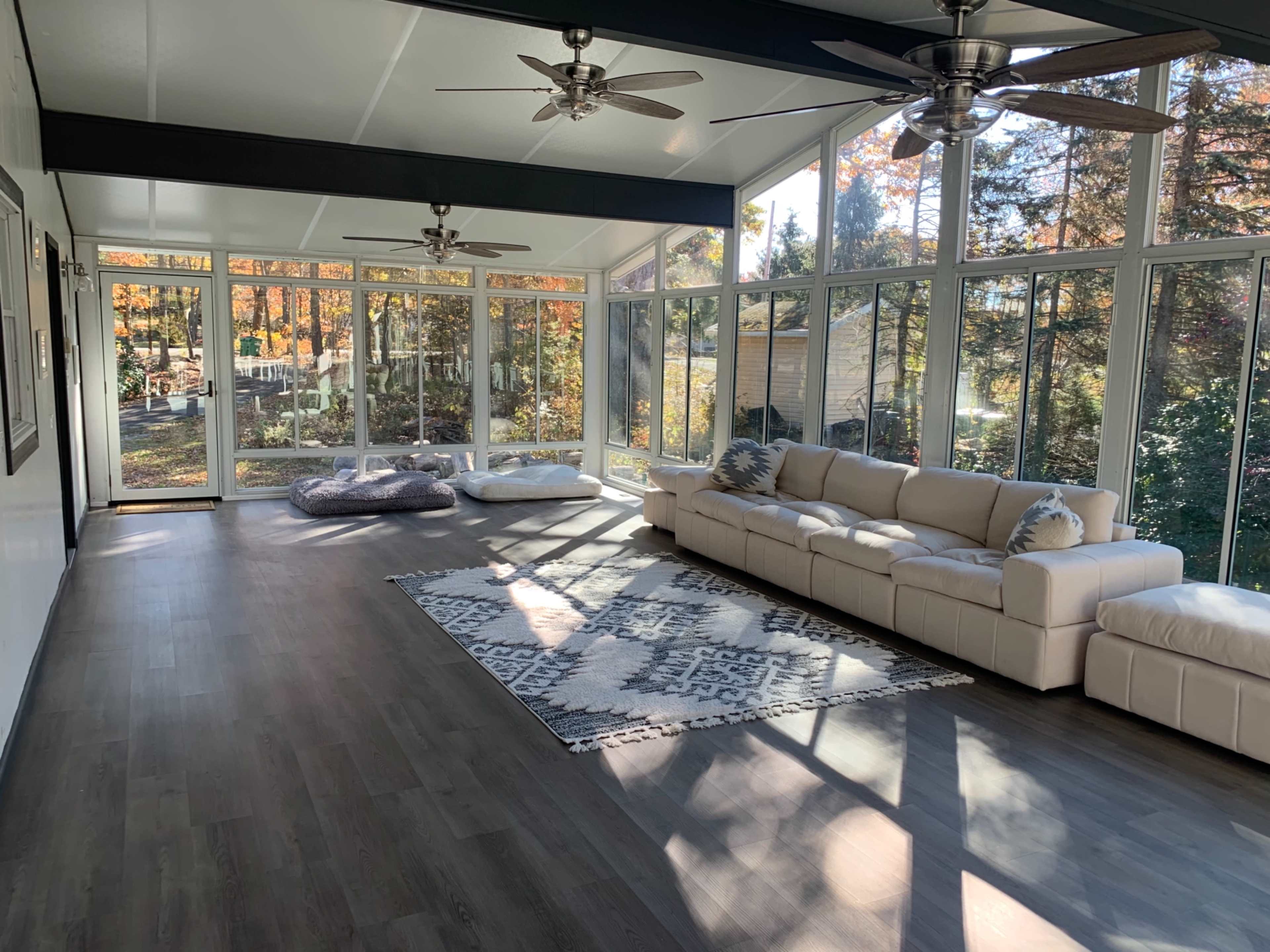 A bright sunroom with large windows, a sectional sofa, a patterned rug, and two ceiling fans.