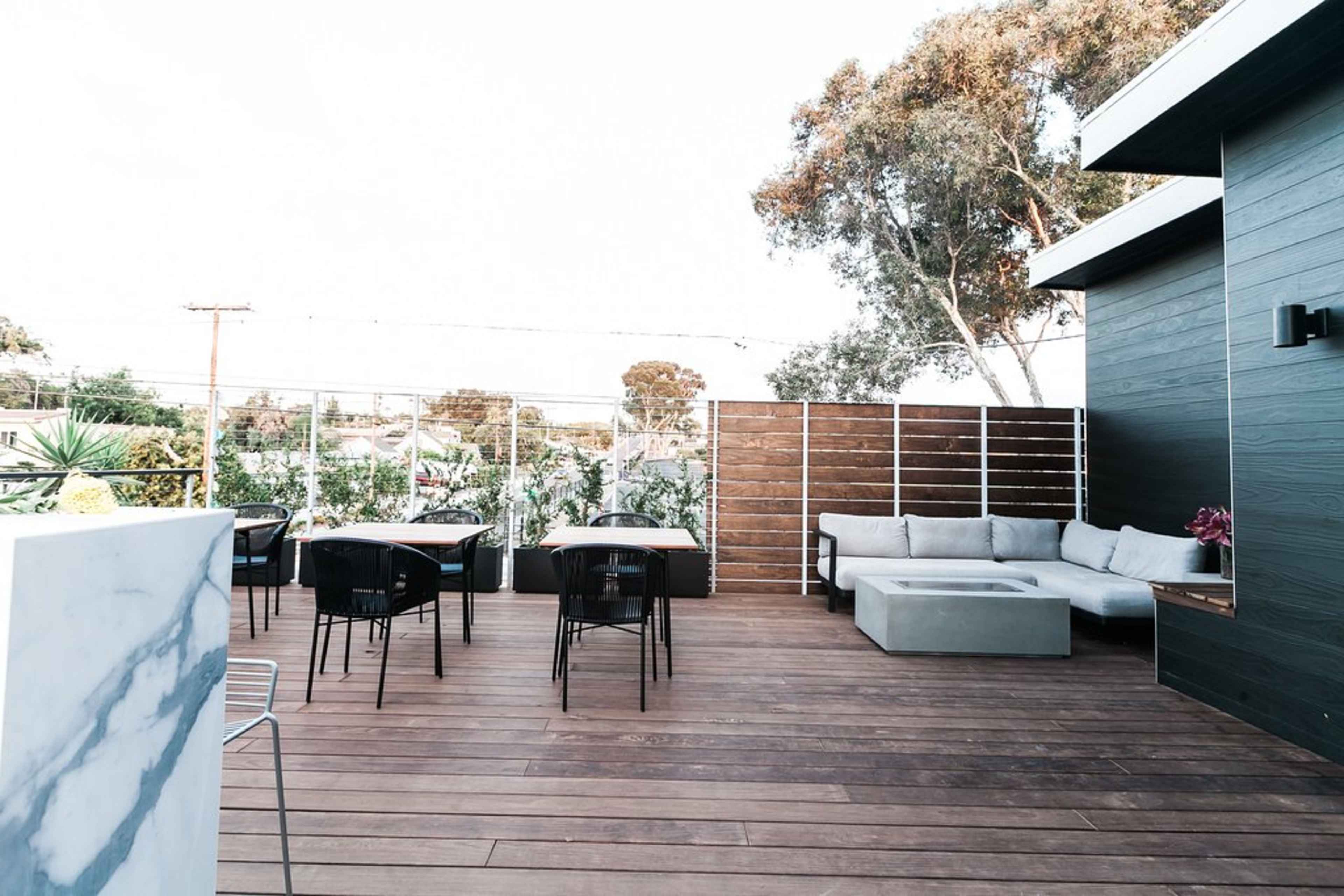 The image shows a modern outdoor patio with wooden decking, a sectional sofa, dining tables, and surrounding greenery.