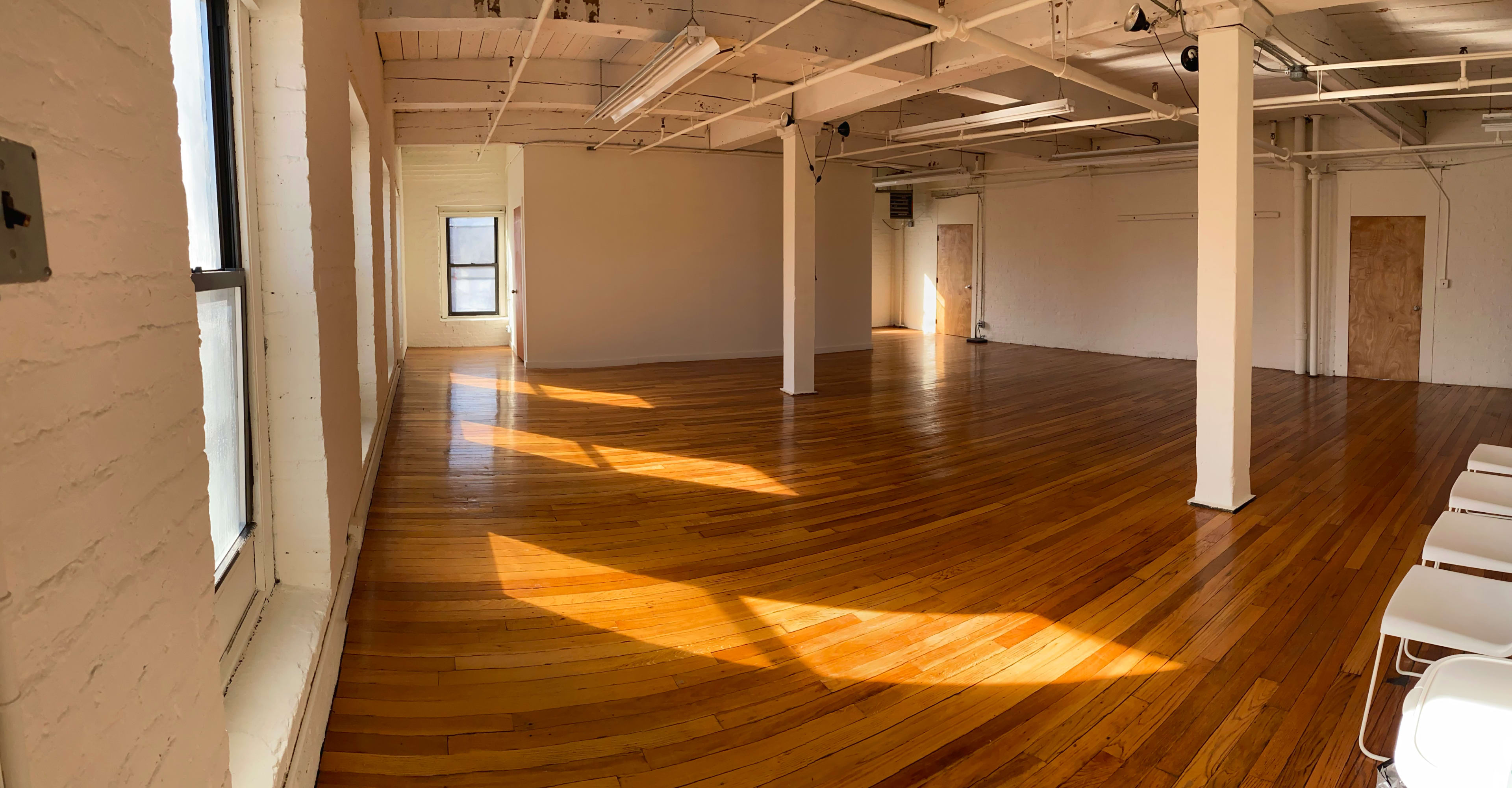 A spacious, empty room with wooden floors and large windows allowing sunlight to illuminate the space.