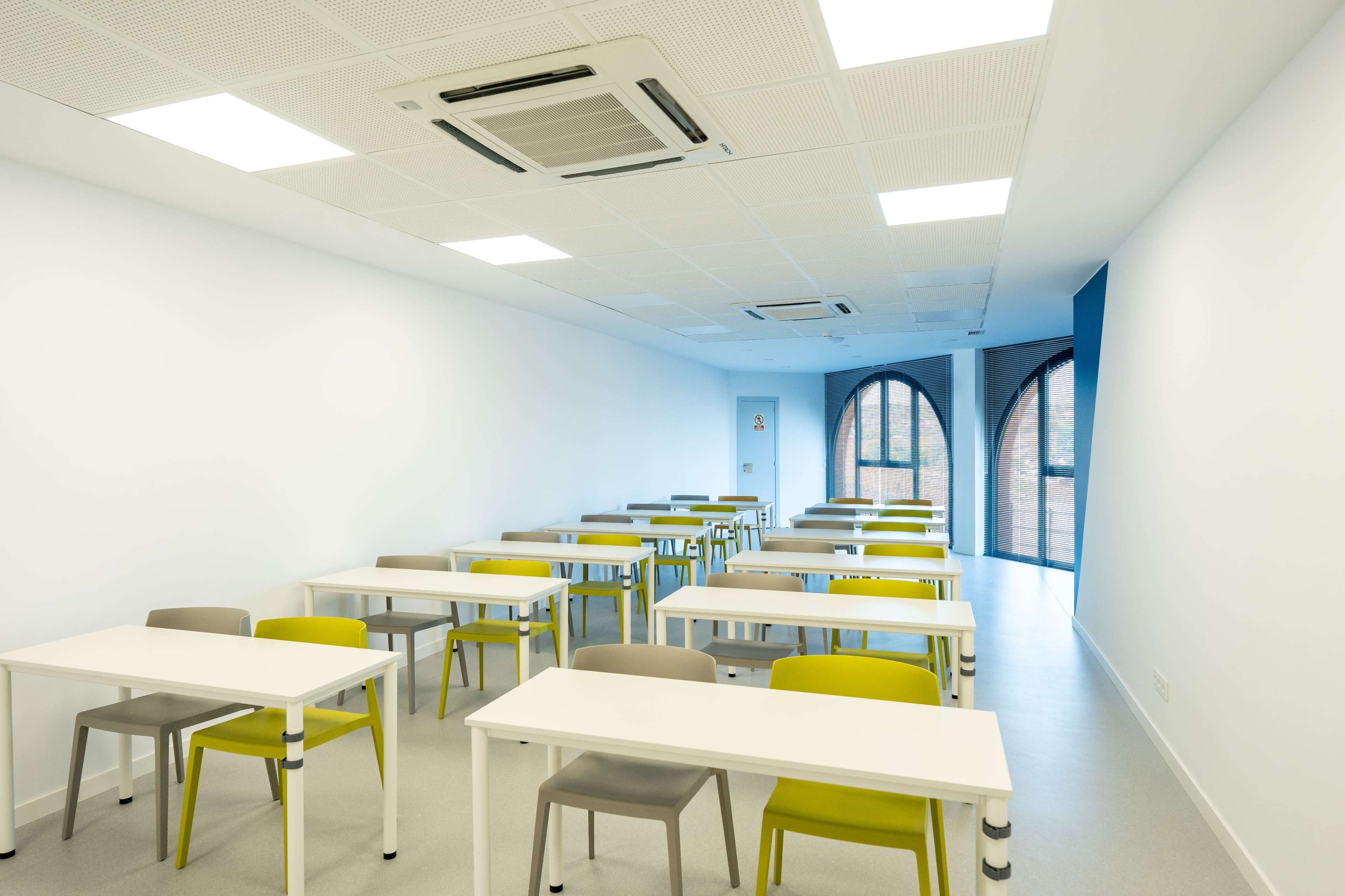 The image shows a modern classroom containing rows of tables and chairs arranged neatly on a light-colored floor.