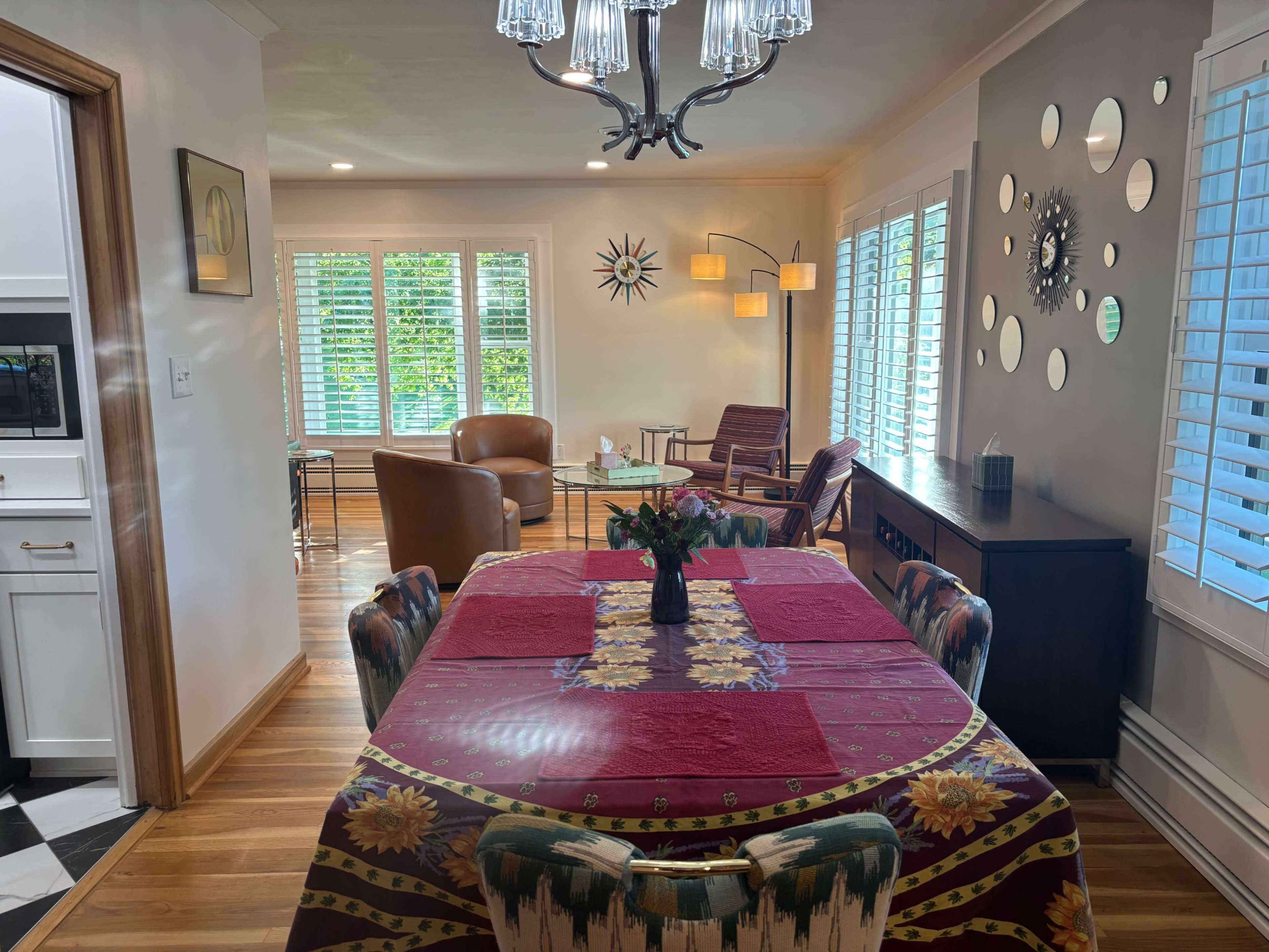 The image shows a dining area with a table set for four, adorned with a patterned tablecloth, and a connected living space featuring two chairs and floor lamps, illuminated by natural light from large windows.