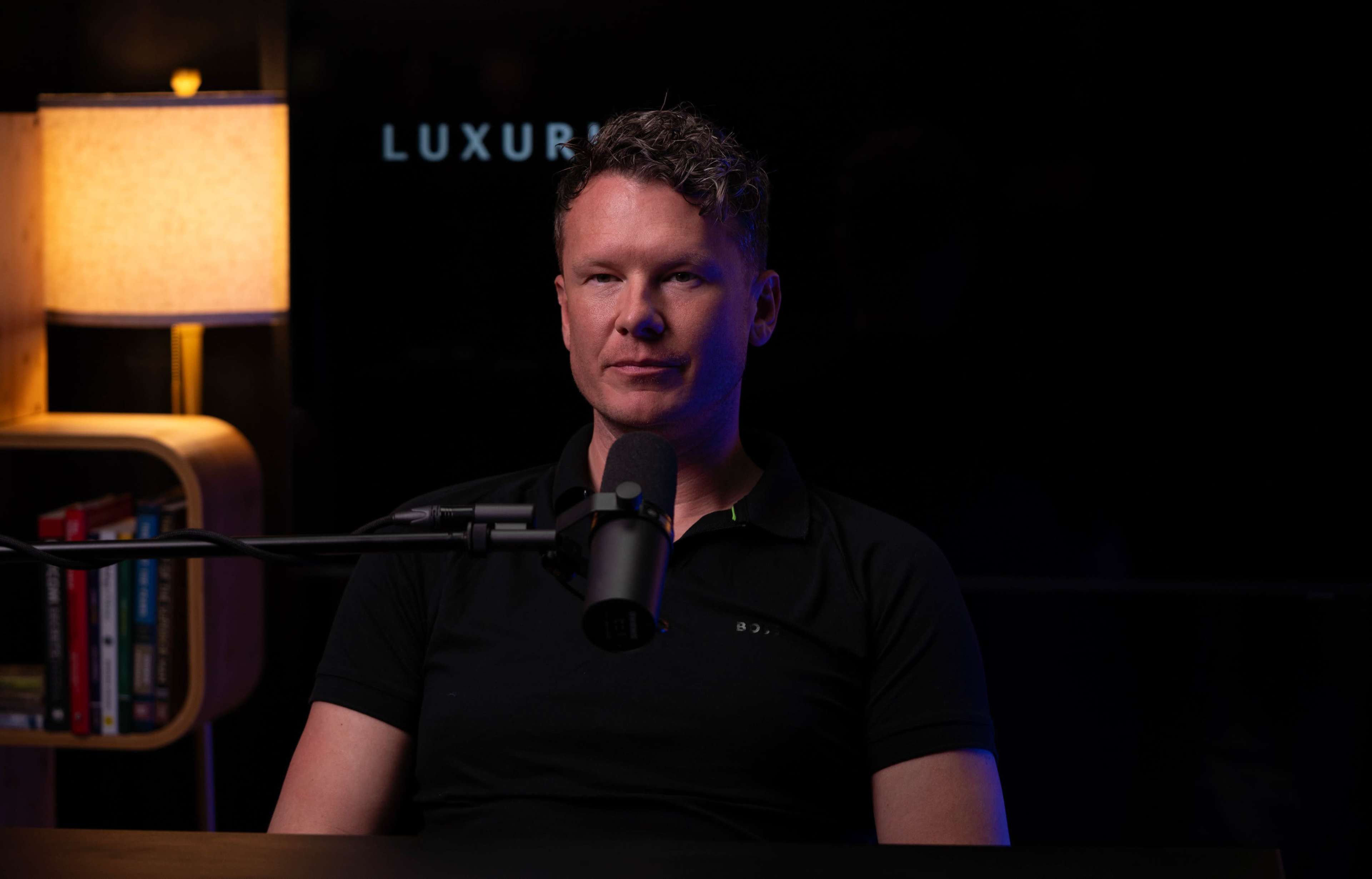 A man sits at a table with a microphone in front of him, illuminated by soft lighting in a dimly lit room.