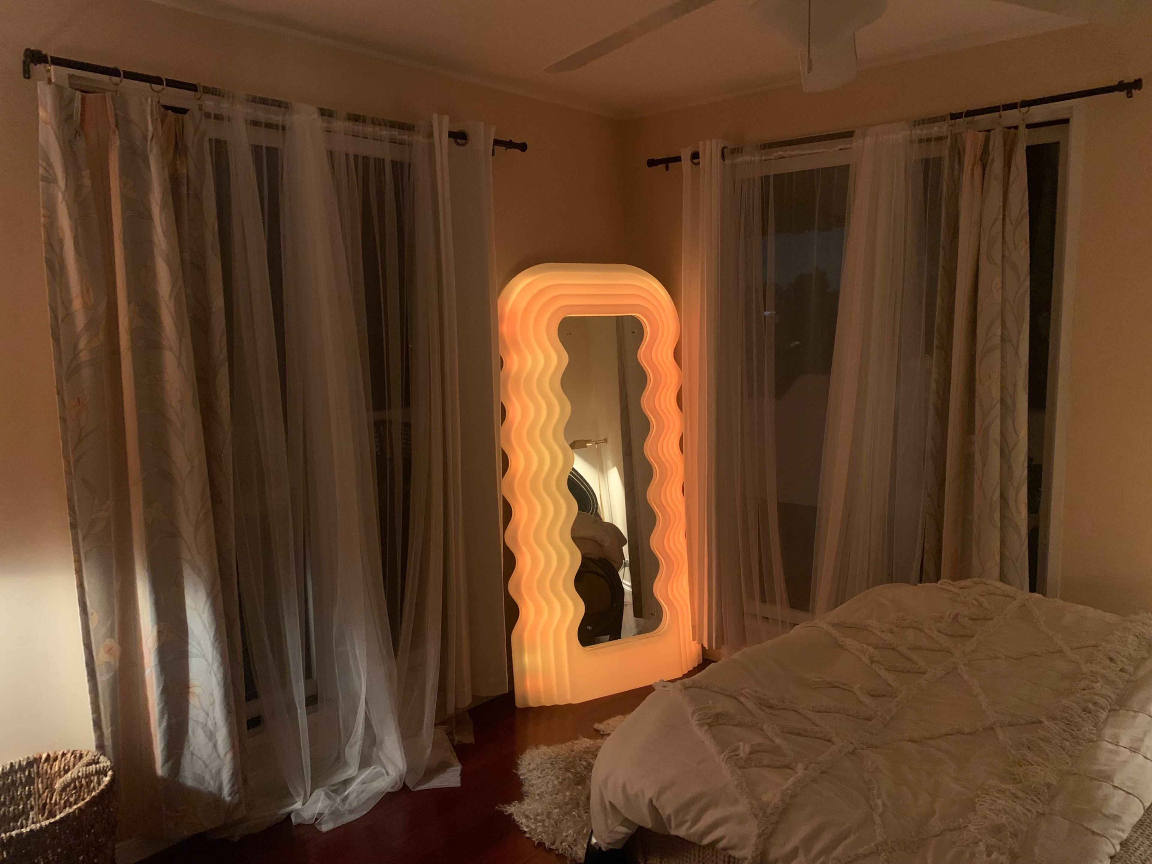 A softly lit wavy mirror stands in a bedroom beside sheer curtains and a bed with a light-colored comforter.