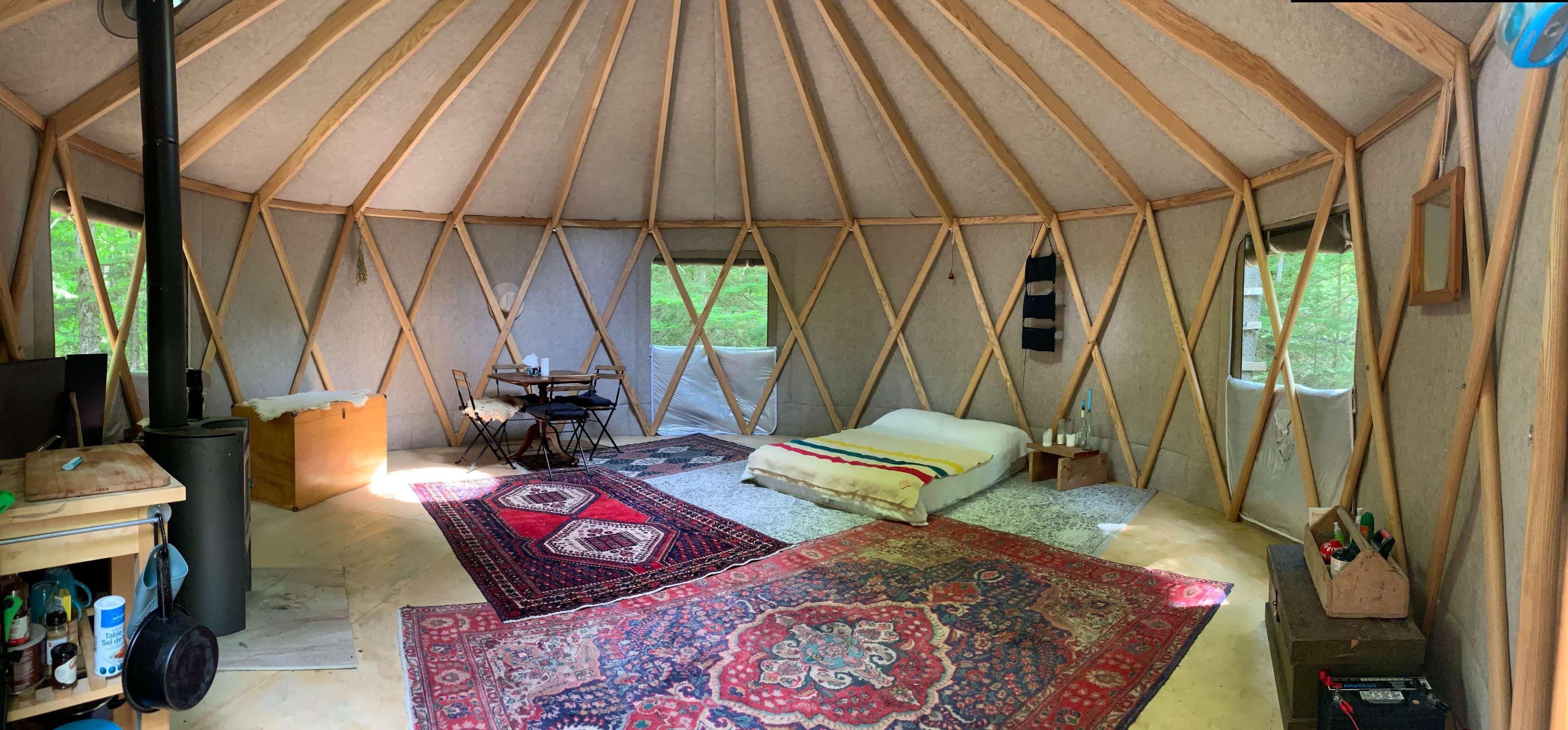 The interior of a yurt features wooden beams, a bed with colorful blankets, a dining table with chairs, and several rugs on the floor.