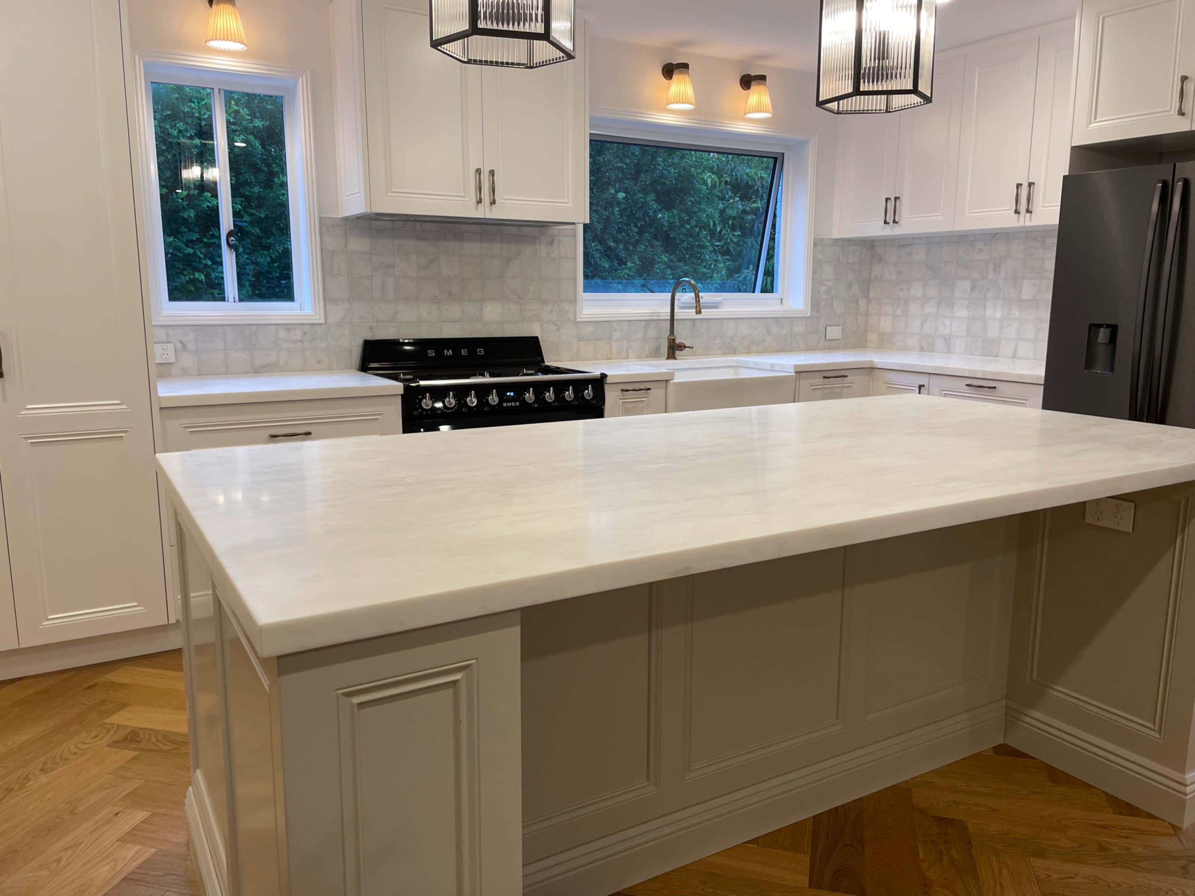 A modern kitchen featuring a large white marble island, stainless steel appliances, and light-colored cabinetry with a herringbone wood floor.