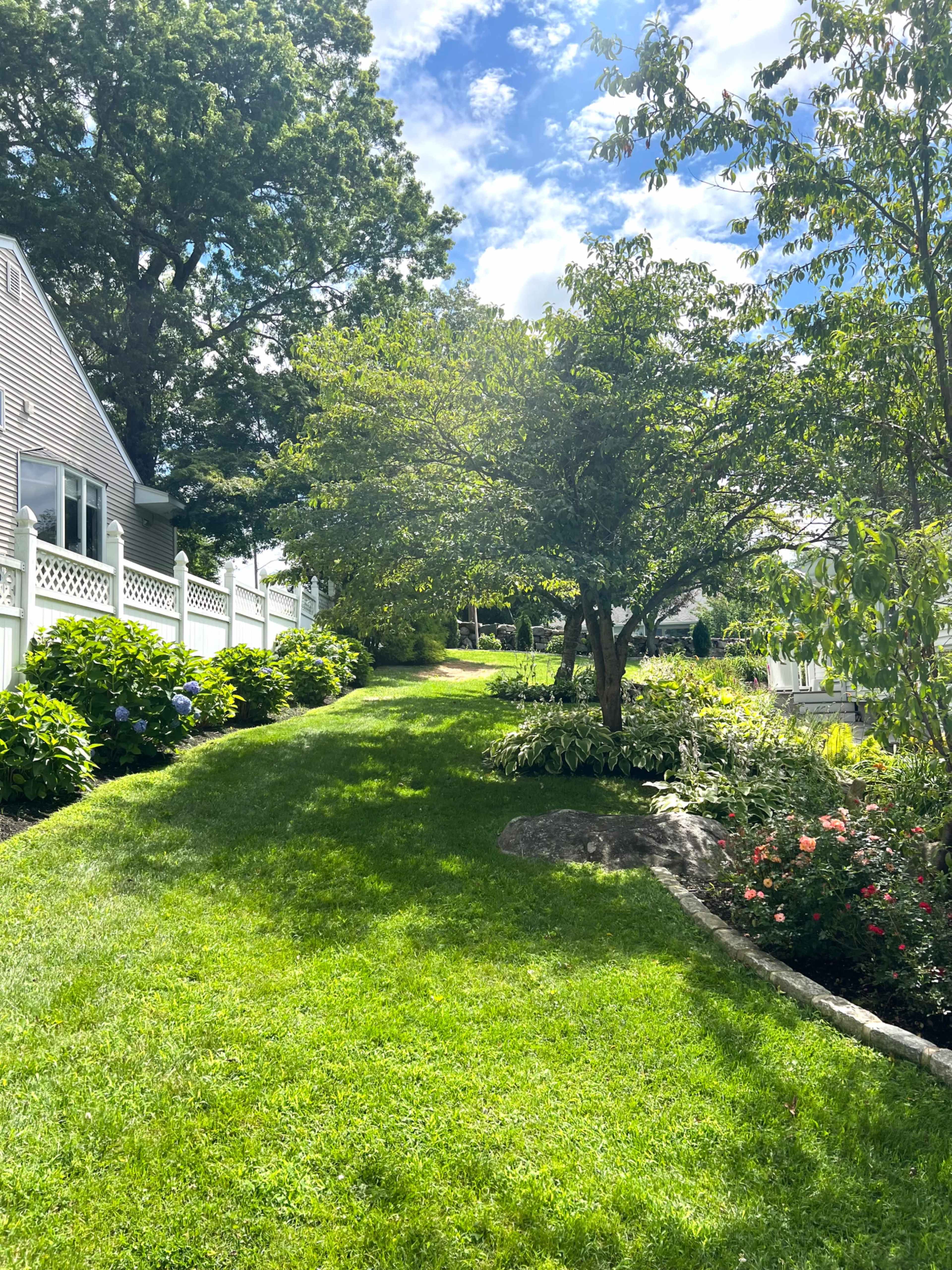 A well-maintained lawn slopes gently upward, bordered by flowering shrubs and trees under a partly cloudy sky.