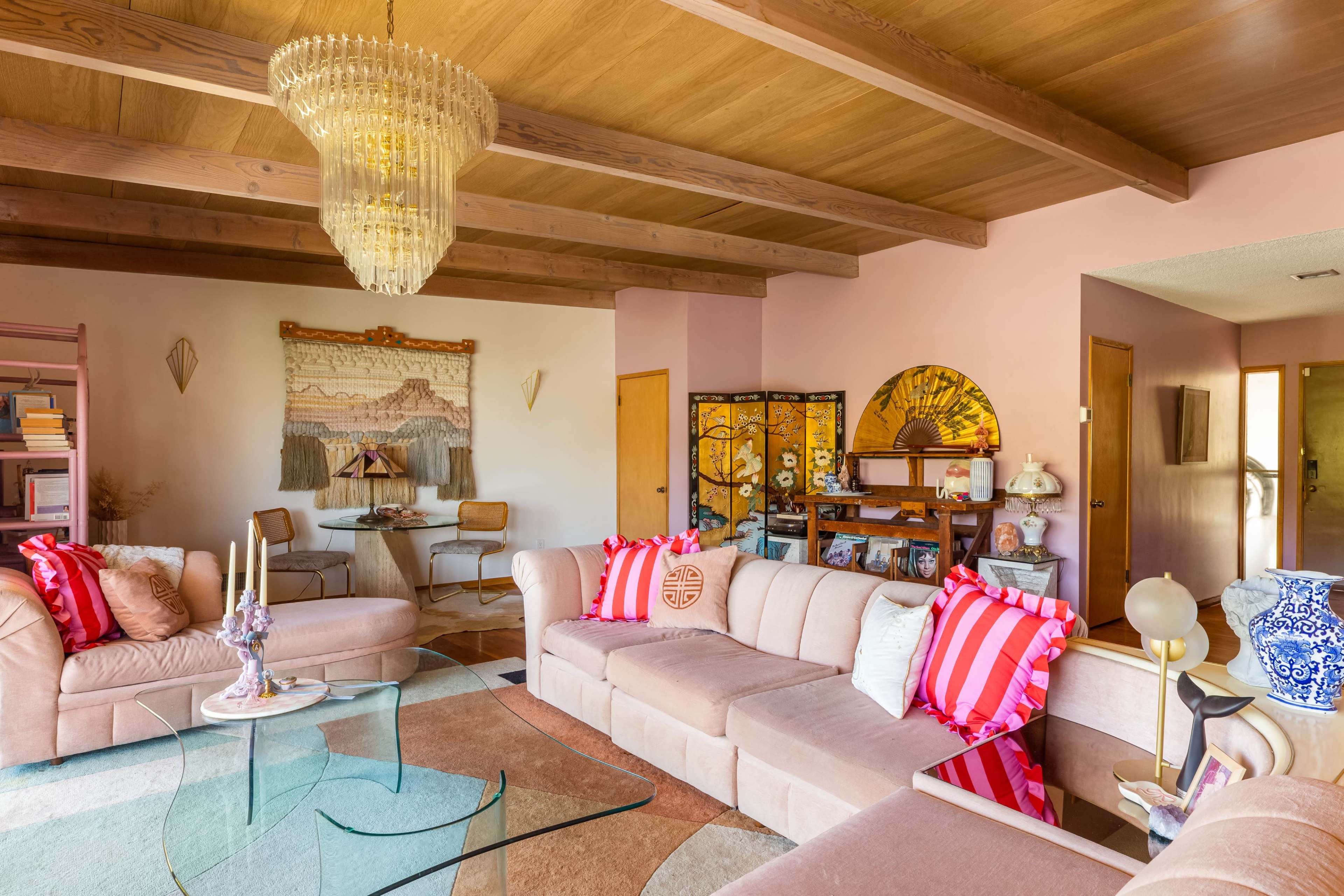 The image shows a cozy living room with a light pink color scheme, featuring a sectional sofa, a glass coffee table, and decorative elements like a large chandelier and artwork on the walls.