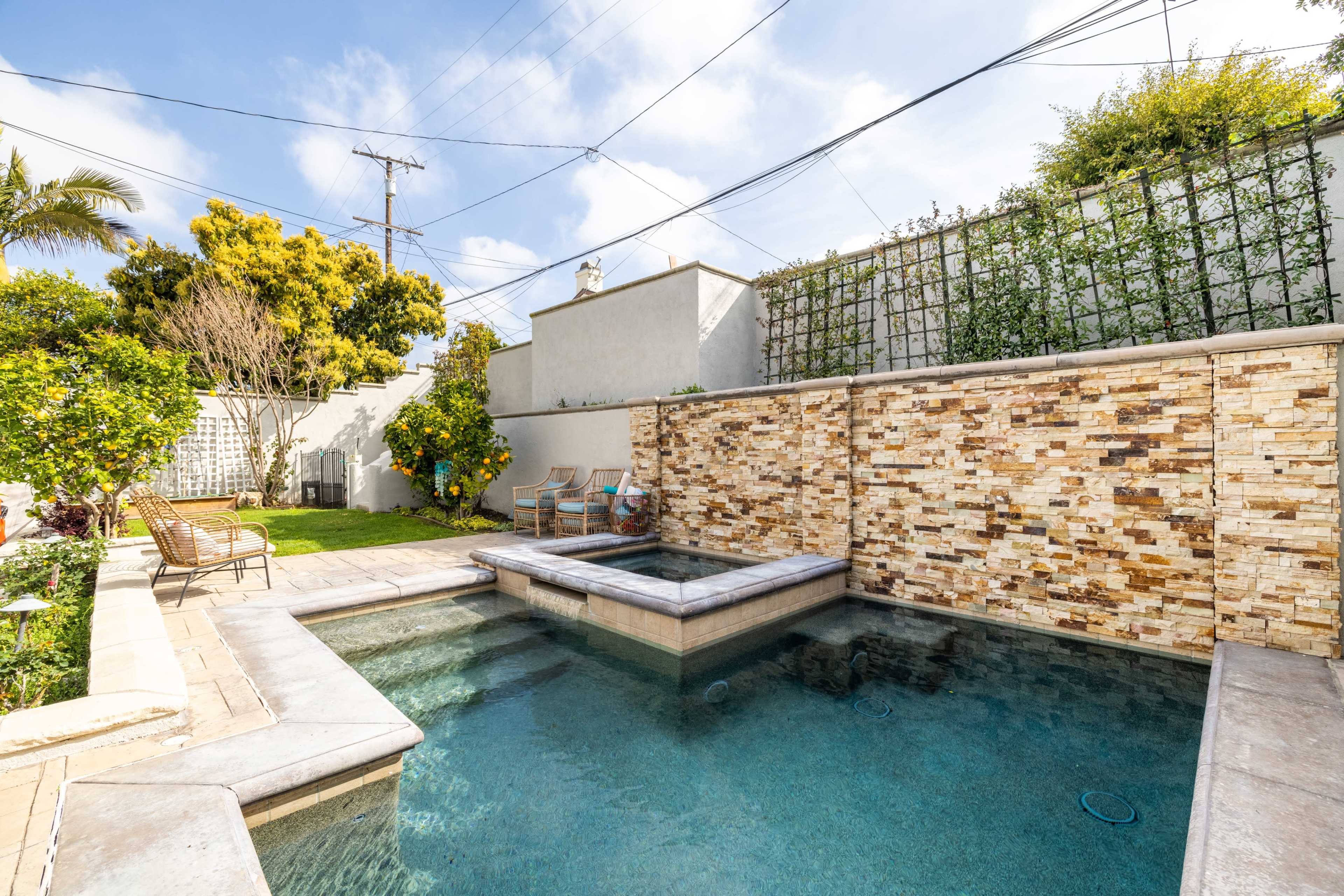 A backyard pool area featuring a stone wall, a spa, and several lounge chairs surrounded by greenery.