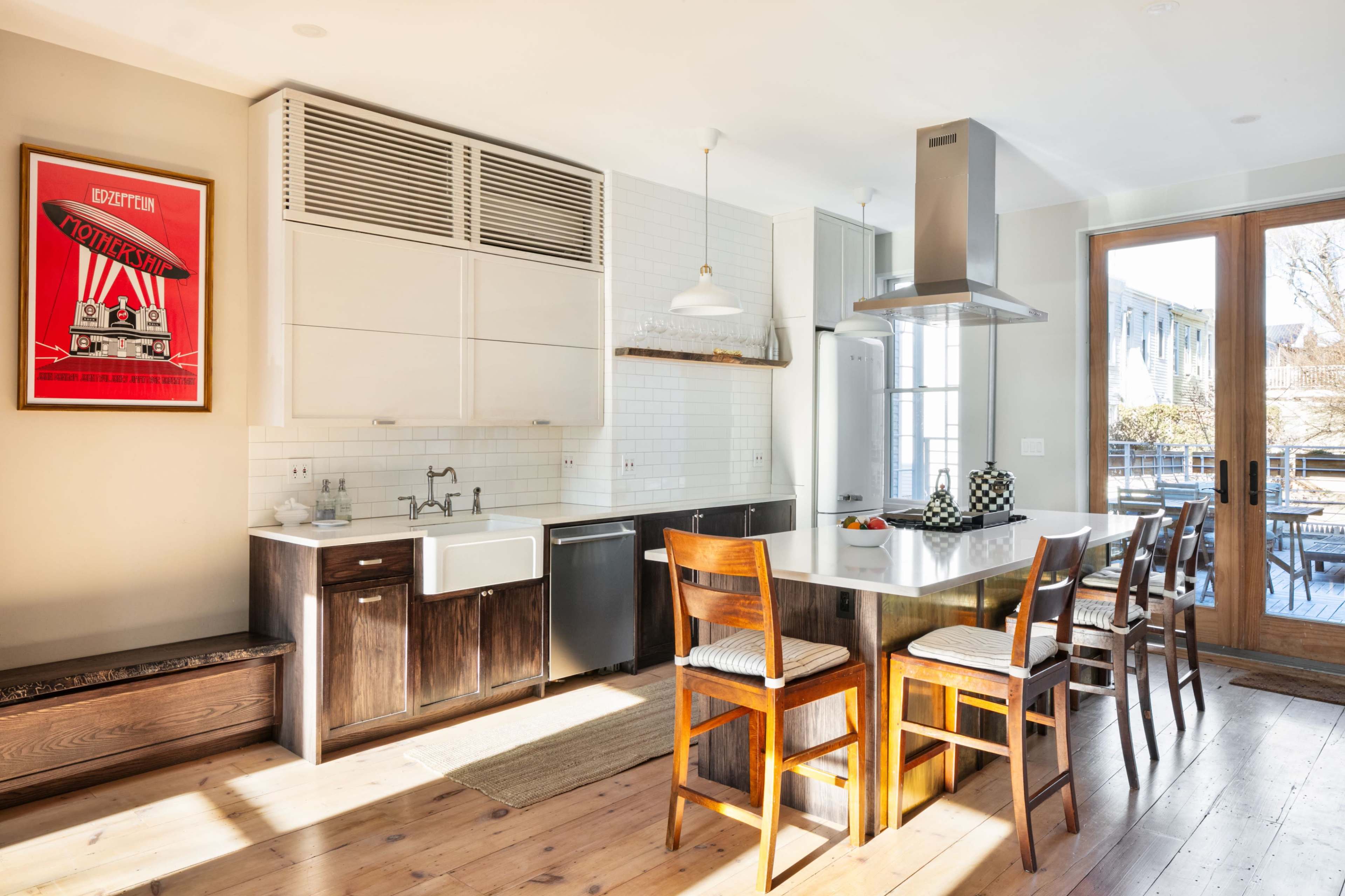 The image shows a bright kitchen featuring a central island with bar stools, modern cabinetry, and large windows leading to an outdoor space.