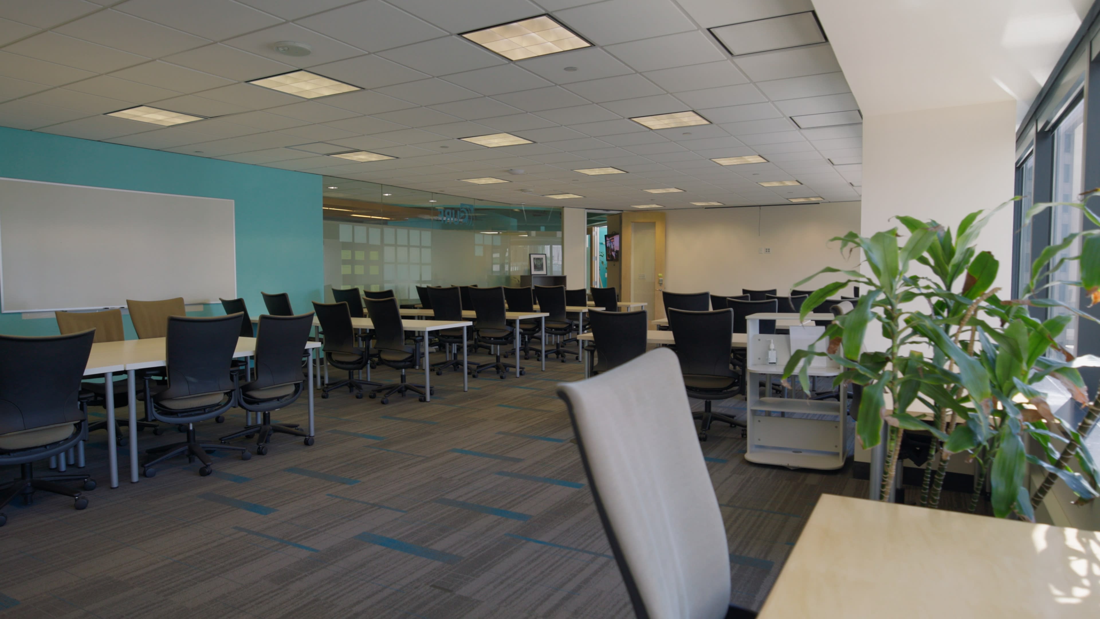 The image shows a spacious office area with multiple empty desks and chairs arranged in rows, featuring a light blue accent wall and large windows.