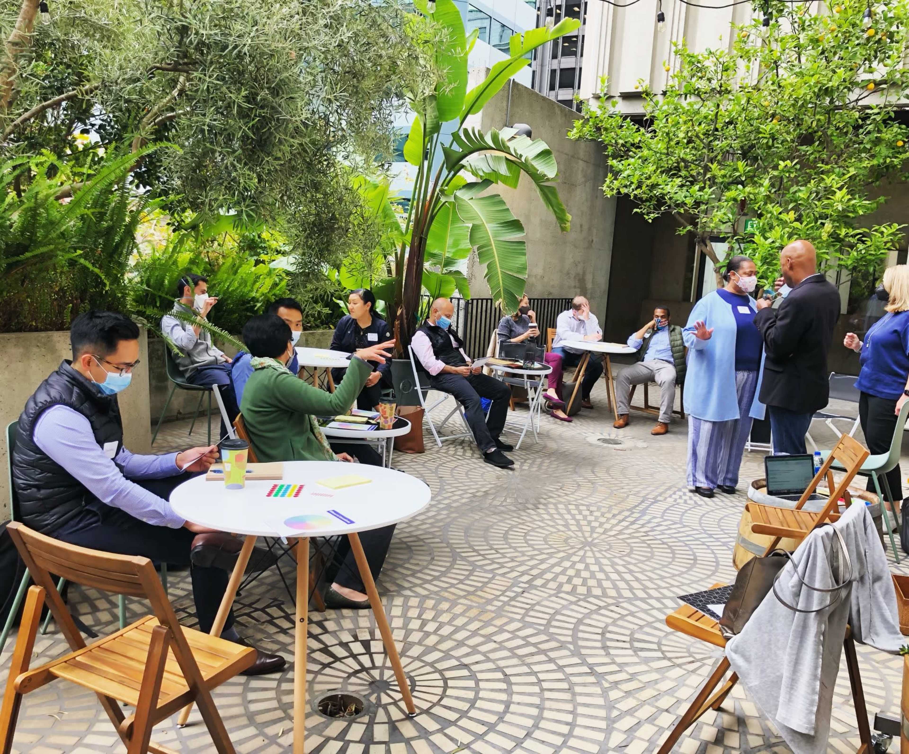 A group of people is engaged in discussions around tables in a landscaped outdoor setting.