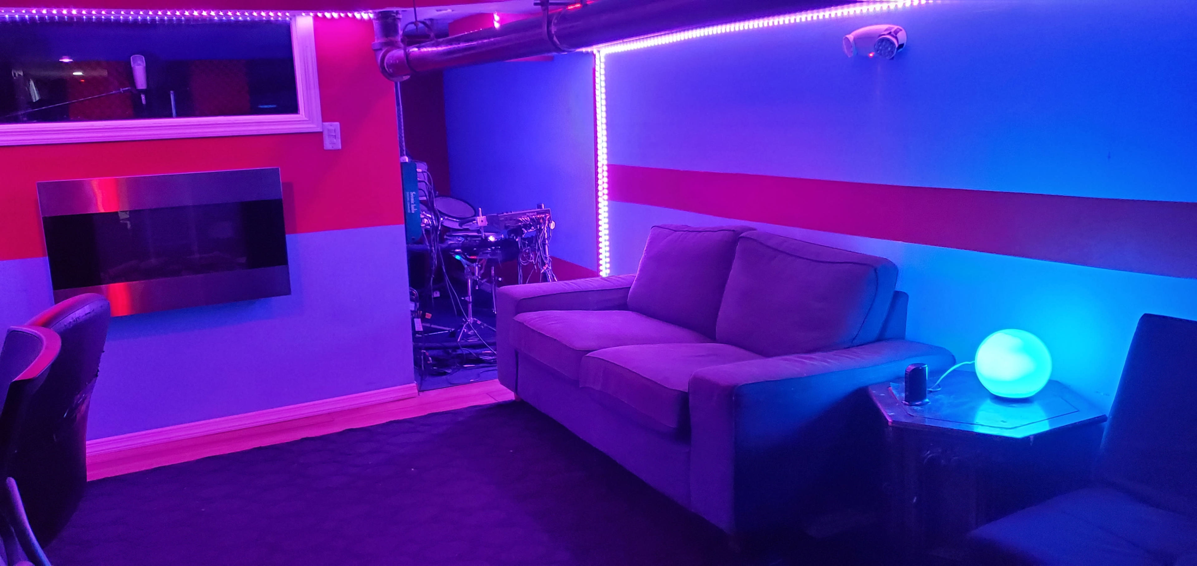 A room with purple and blue lighting, a gray couch, a small table, and a drum set in the background.