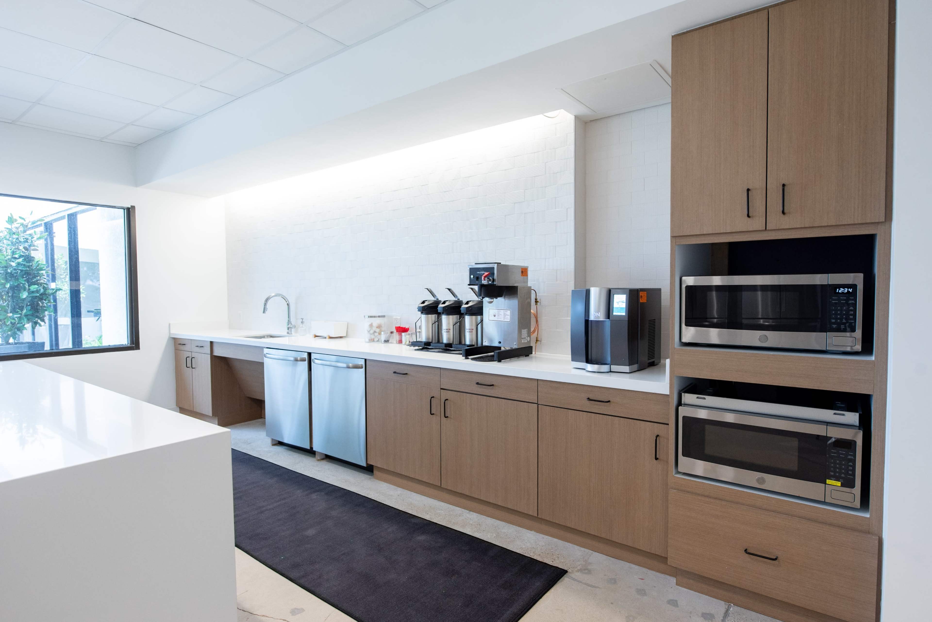 A modern kitchen with a white countertop, wooden cabinetry, a coffee machine, a water dispenser, and two microwaves.