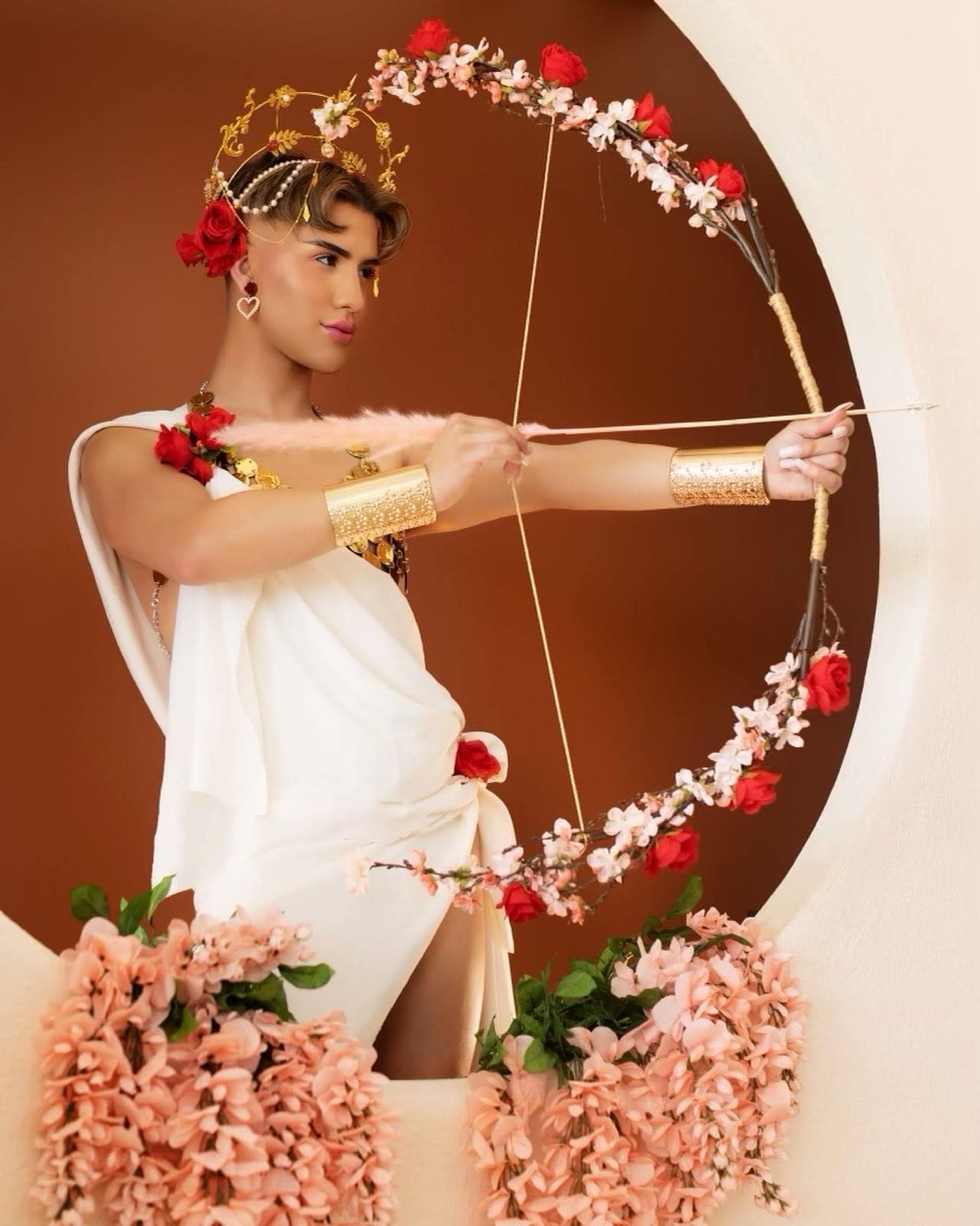 A person dressed in a classical-style outfit, adorned with flowers and accessories, aims an ornate bow decorated with blossoms.