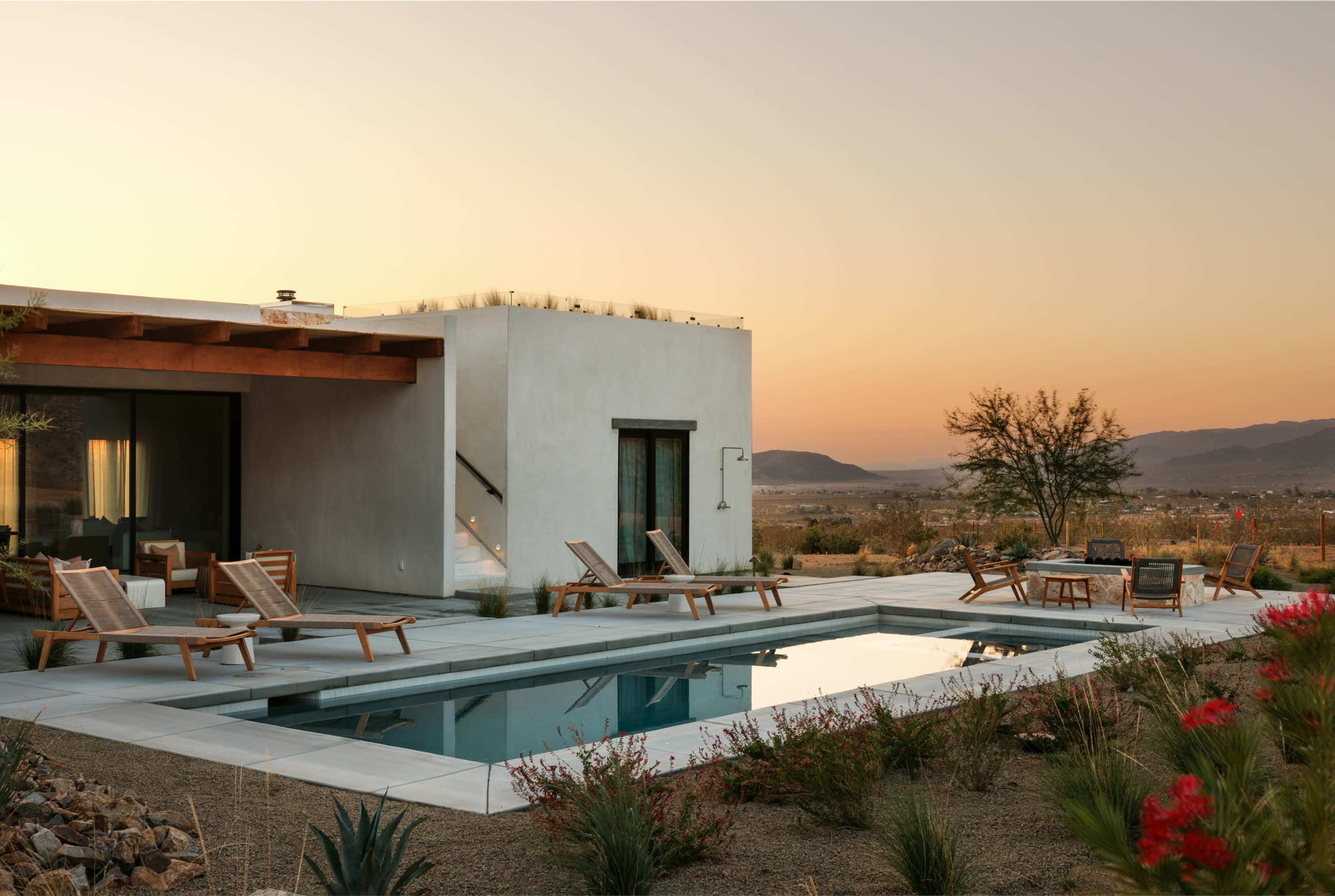 A modern single-story house with large windows overlooks a swimming pool and arid landscape at sunset.