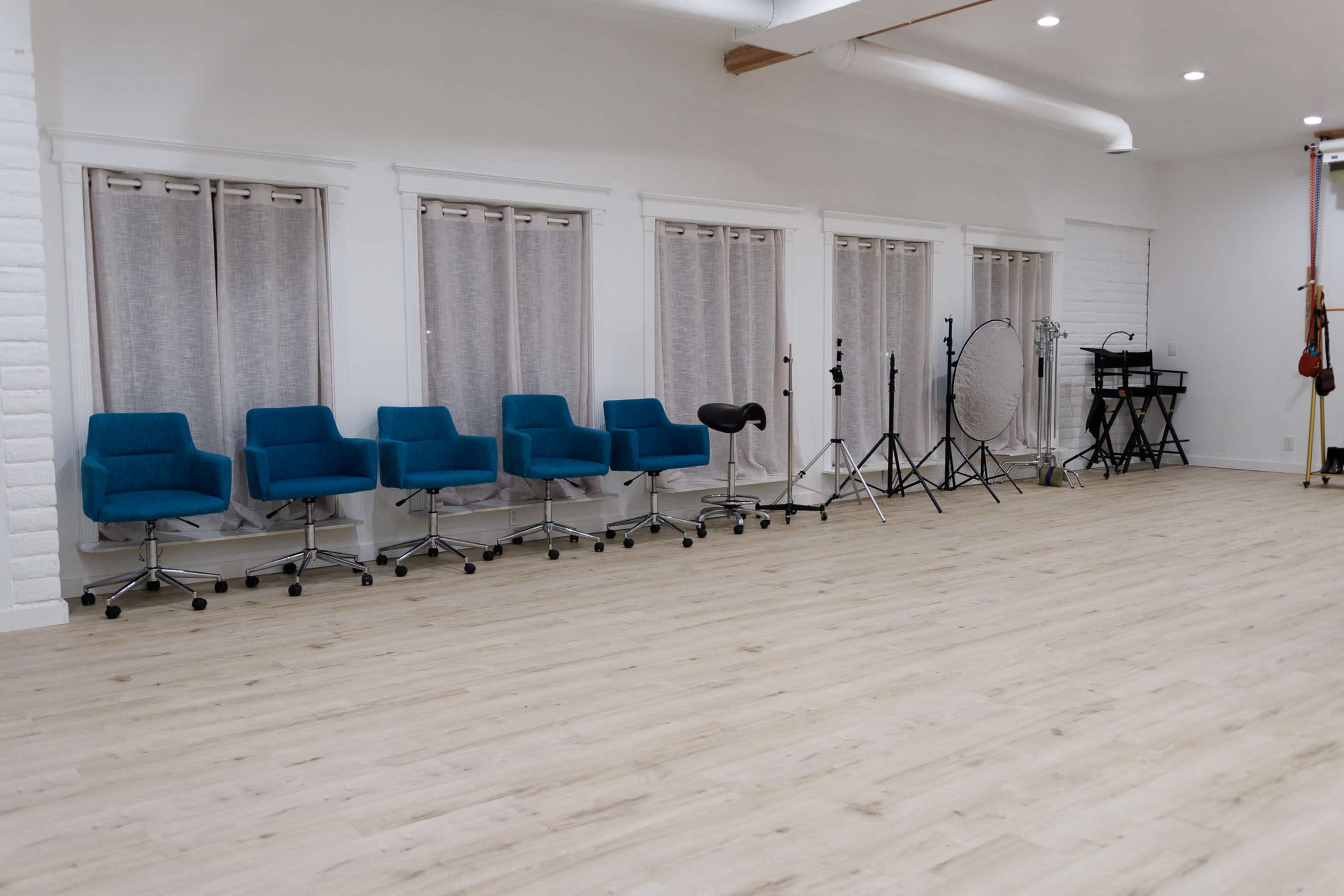 The image shows a spacious room featuring several blue chairs lined up against white walls with large windows and a variety of photography equipment positioned nearby.