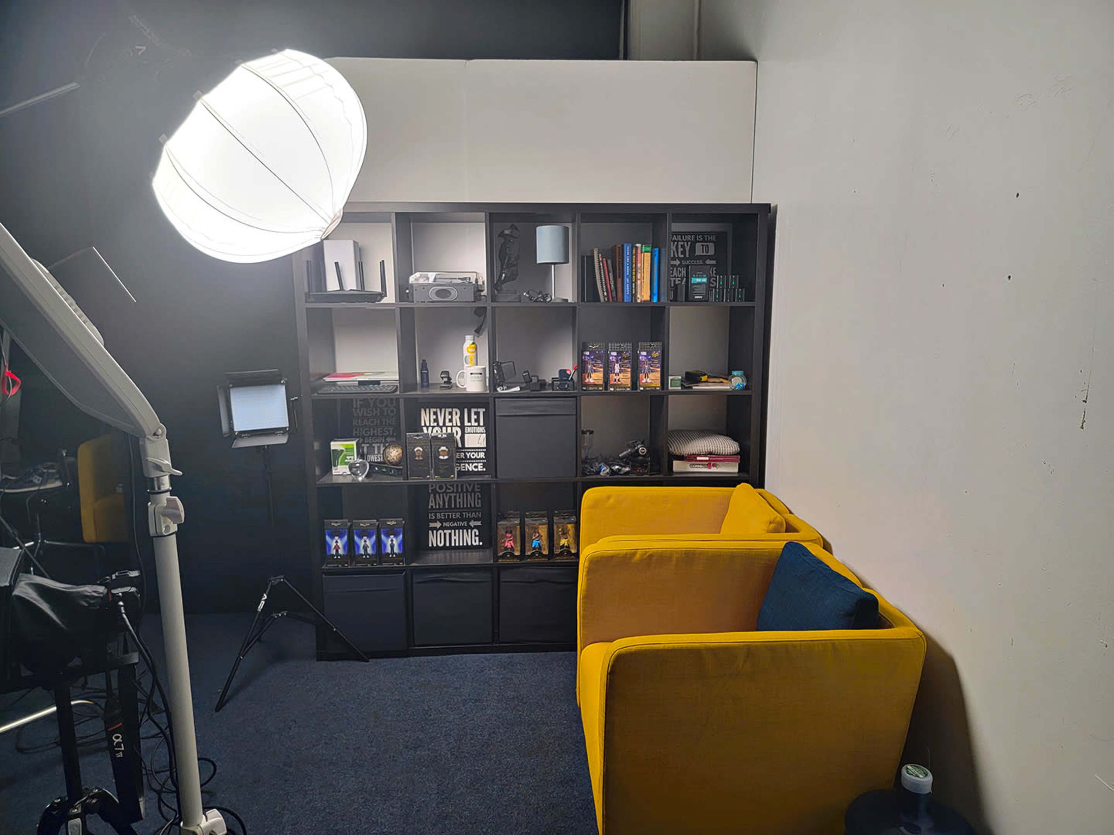 The image shows a neatly arranged studio space with two yellow chairs, a bookshelf filled with various items, and a lighting setup in the corner.