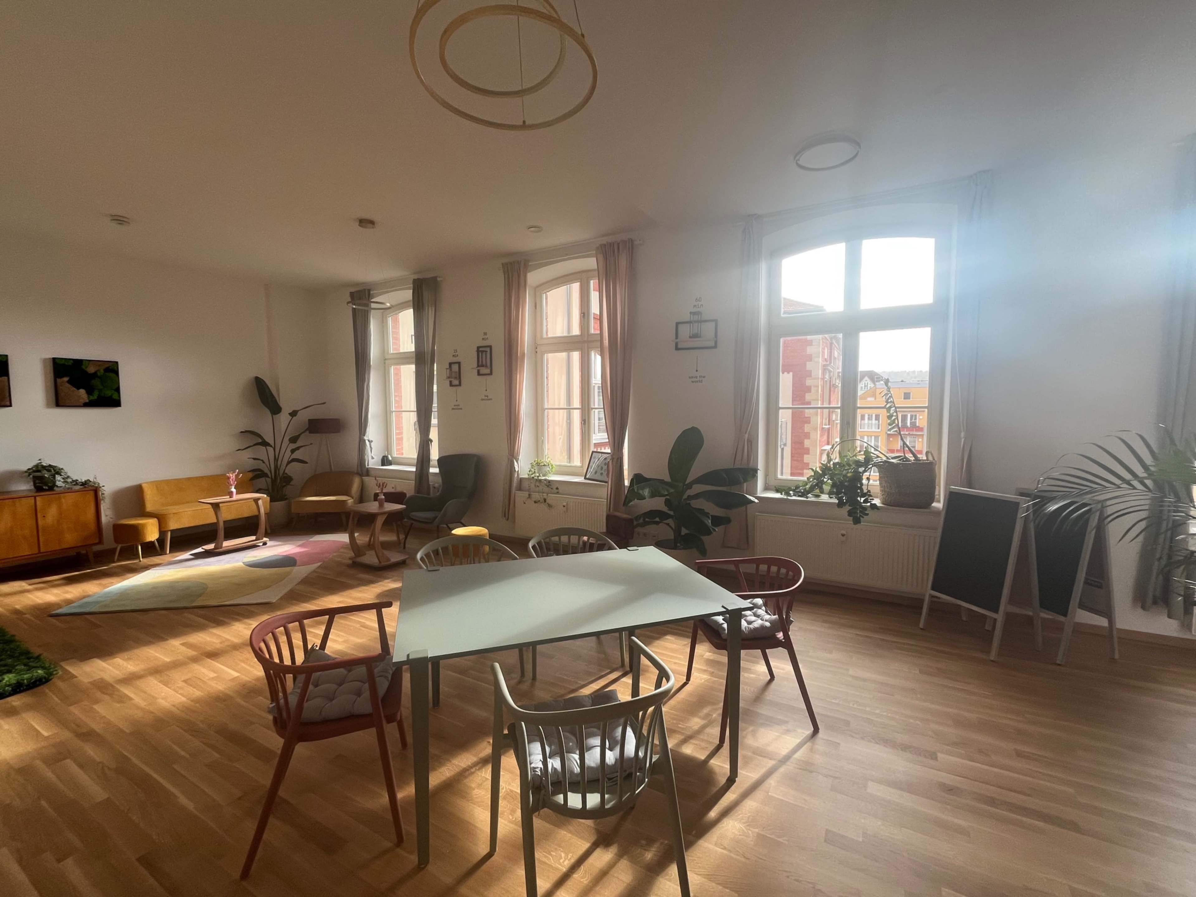 The image shows a bright, modern room with a table surrounded by colorful chairs, large windows letting in natural light, and plants placed around the space.