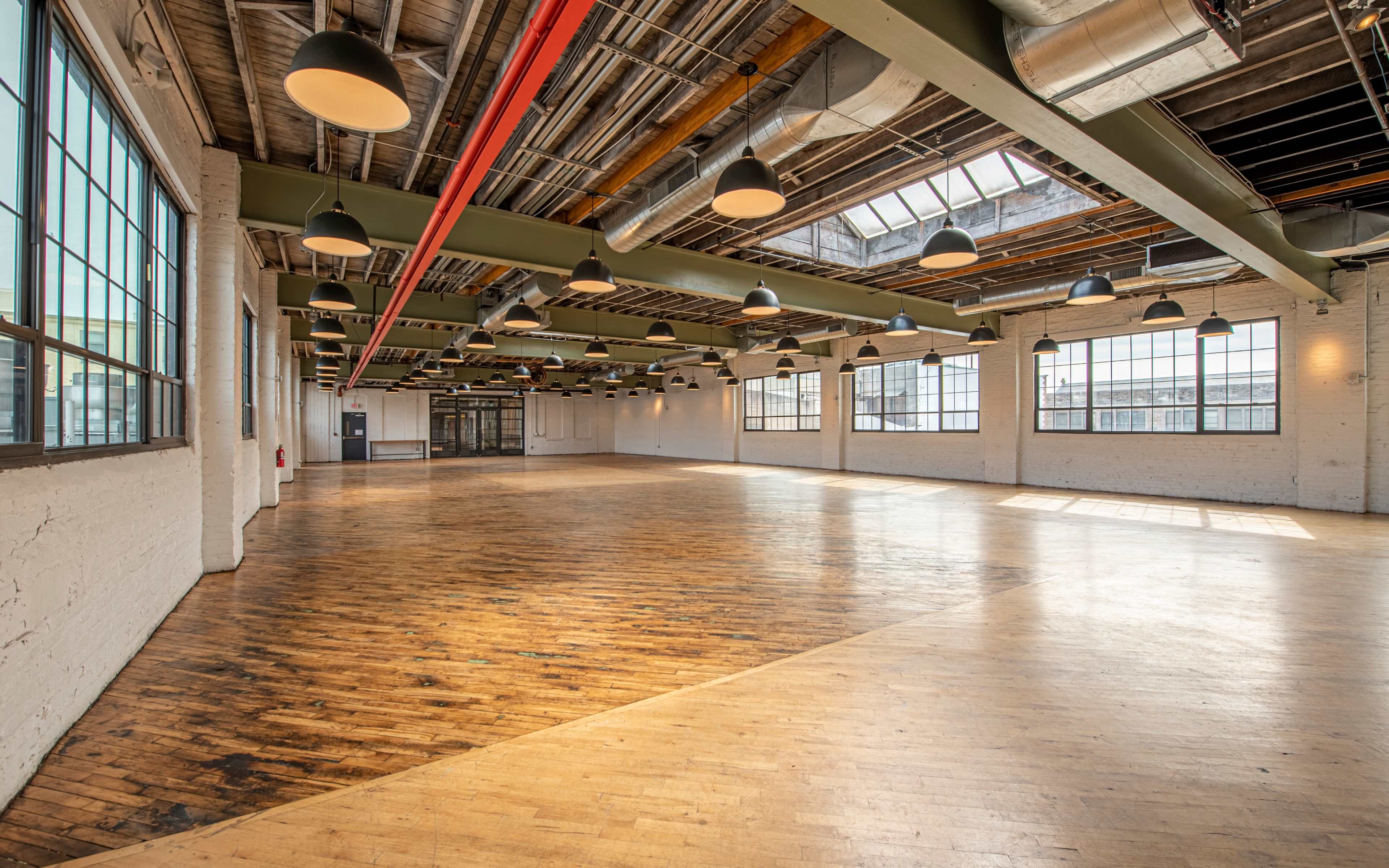A spacious, empty industrial-style room with wooden floors, large windows, and hanging light fixtures.