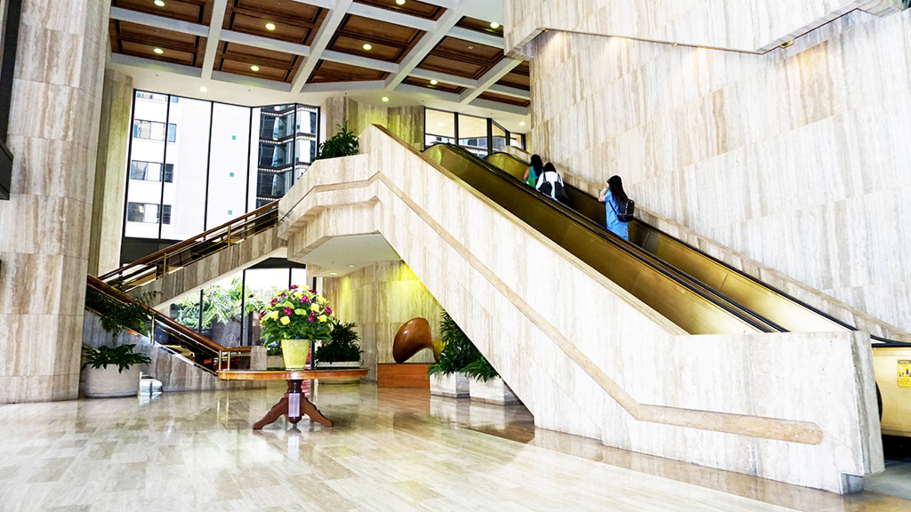A spacious lobby with marble walls and two escalators leading to an upper level, flanked by large plants and a floral arrangement in the foreground.