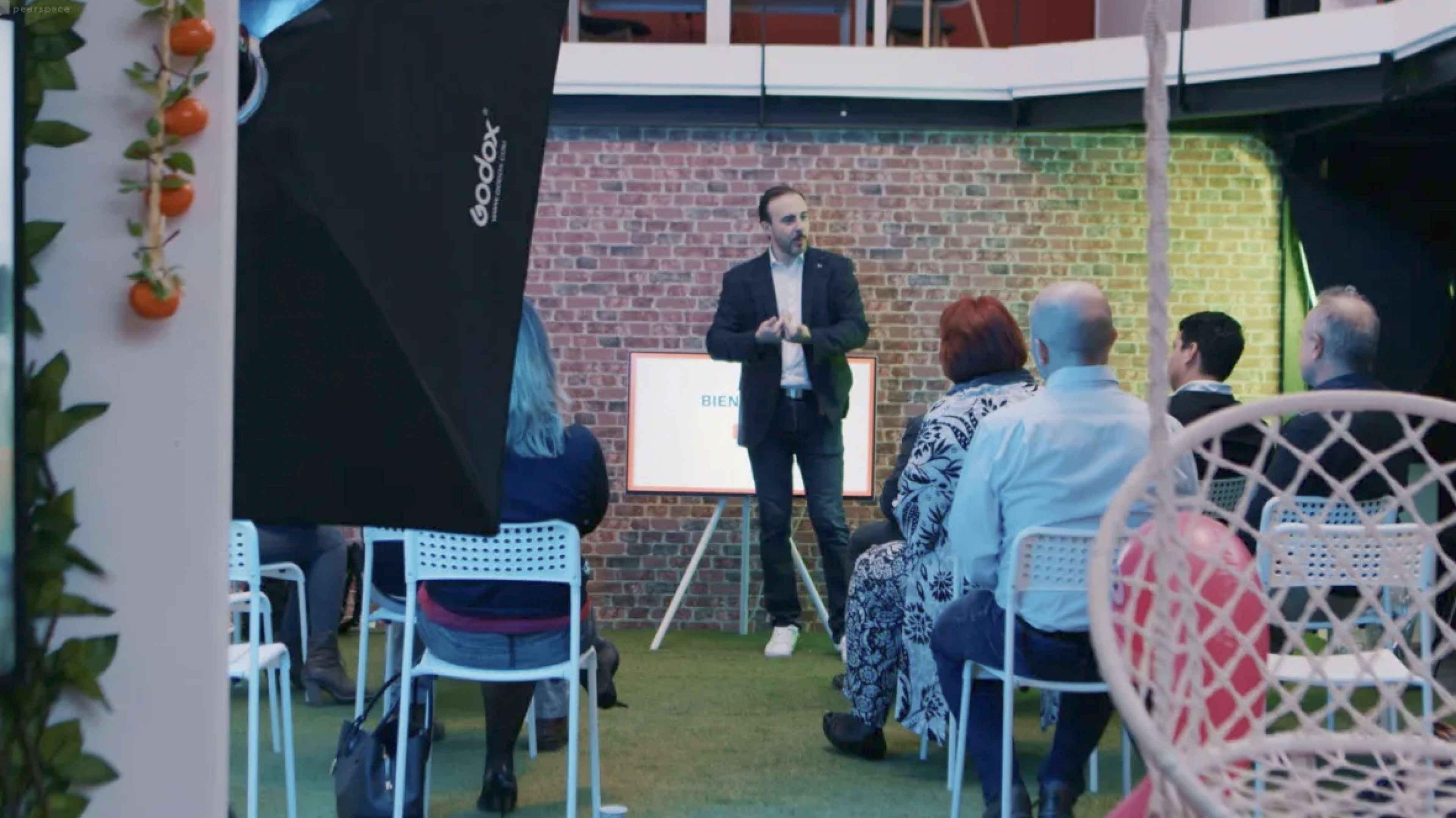 A speaker stands in front of a seated audience in a space decorated with a brick wall and greenery.