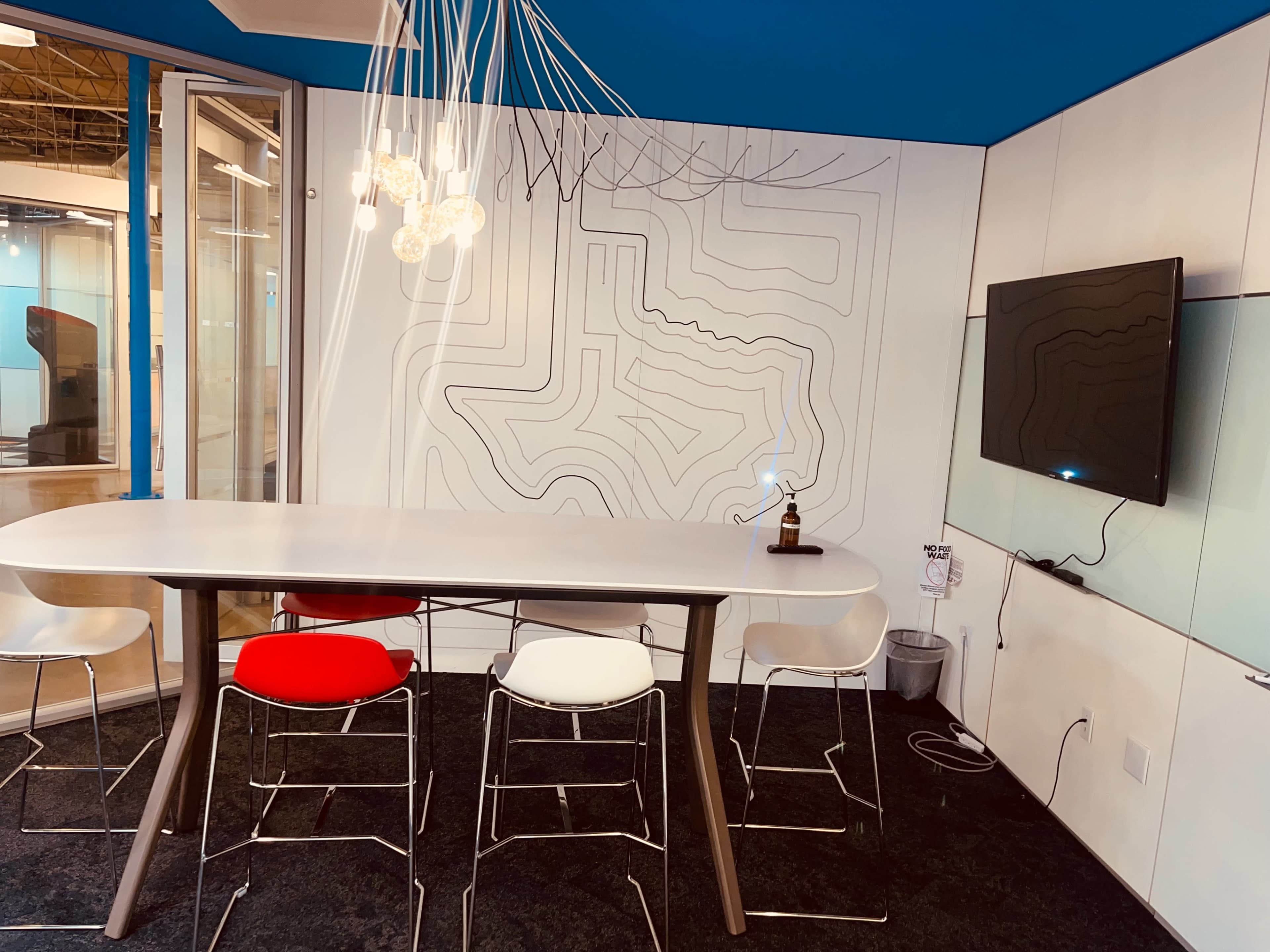 A modern meeting room features a sleek table with white, red, and black chairs, a television on the wall, and a decorative light fixture above.