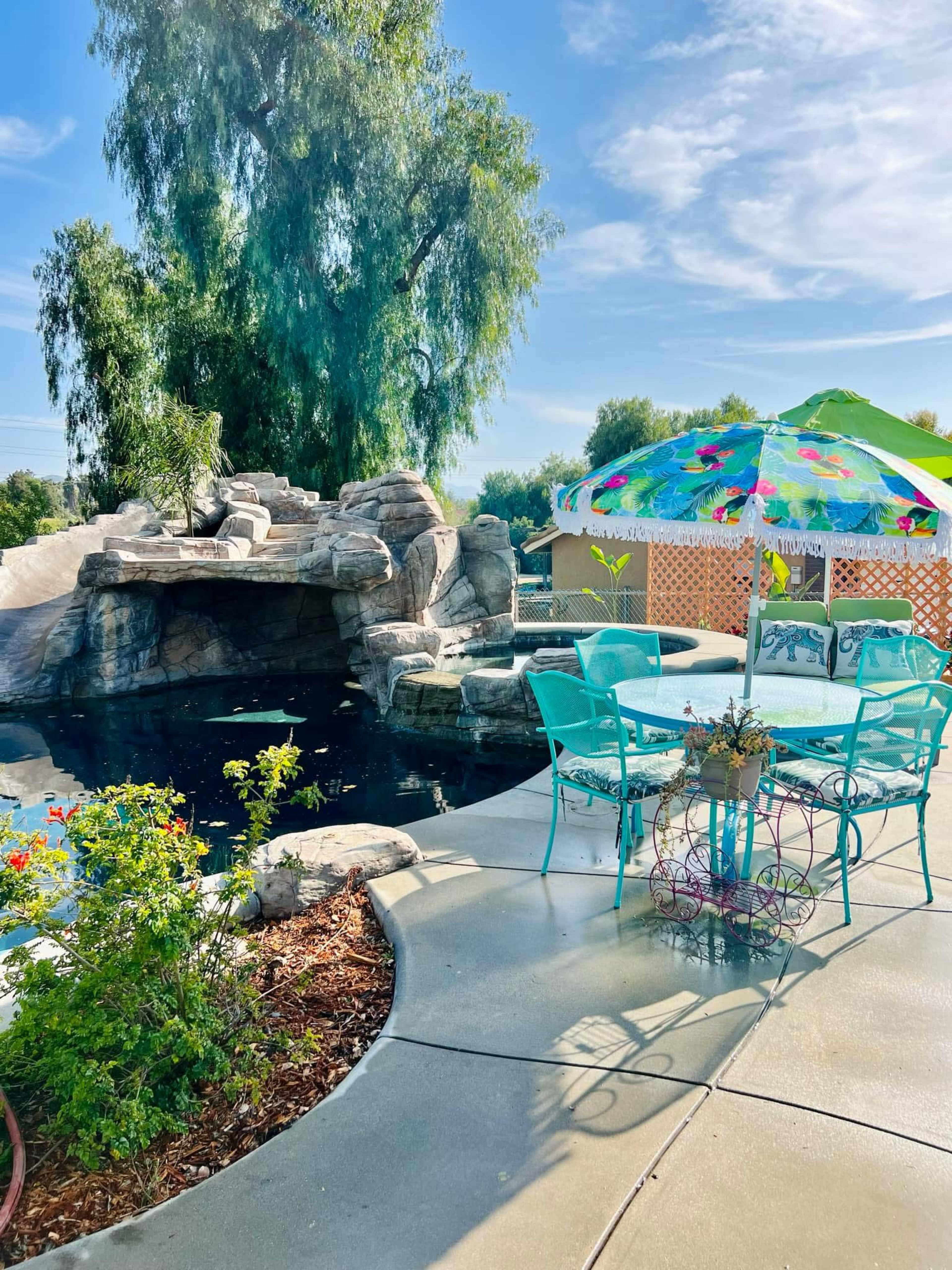 The image shows a backyard pool area featuring a rock formation, a table with four turquoise chairs, and a colorful umbrella, surrounded by greenery and flowers.