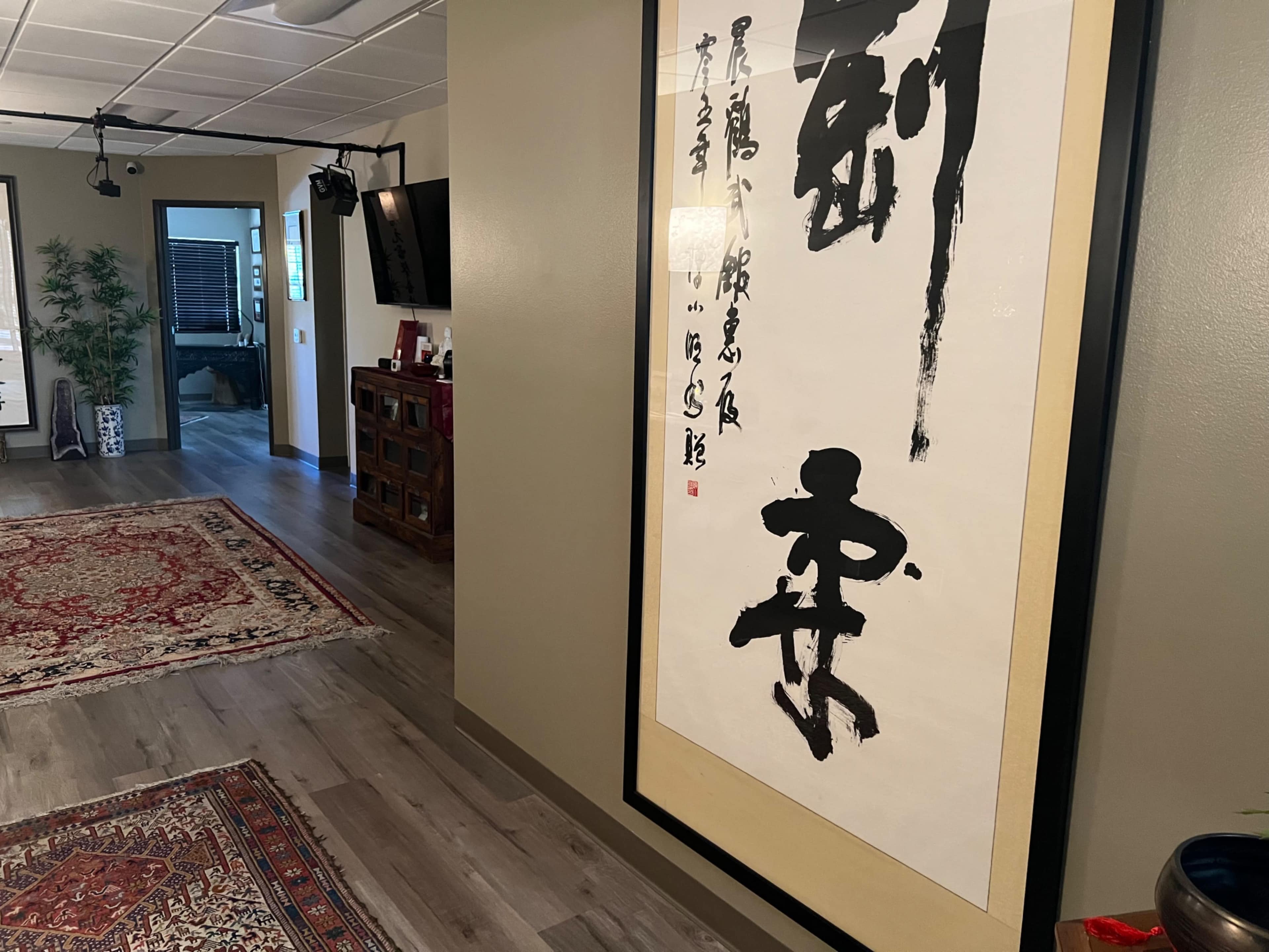 The image shows a hallway with a framed piece of calligraphy hanging on the wall, alongside a patterned rug and a wooden cabinet.