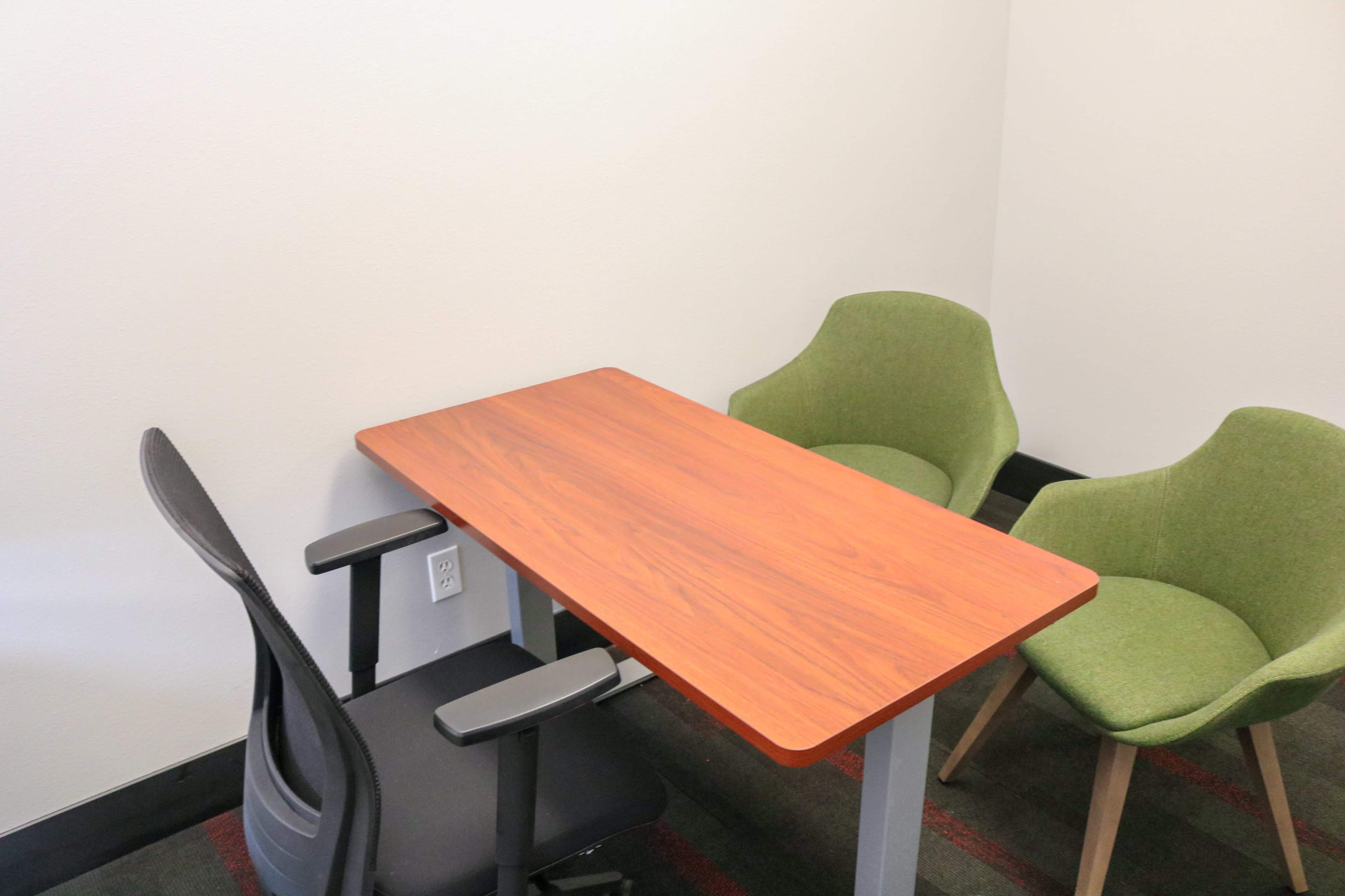 The image shows a small meeting room with a rectangular wooden table and three chairs, two of which are green and one black.