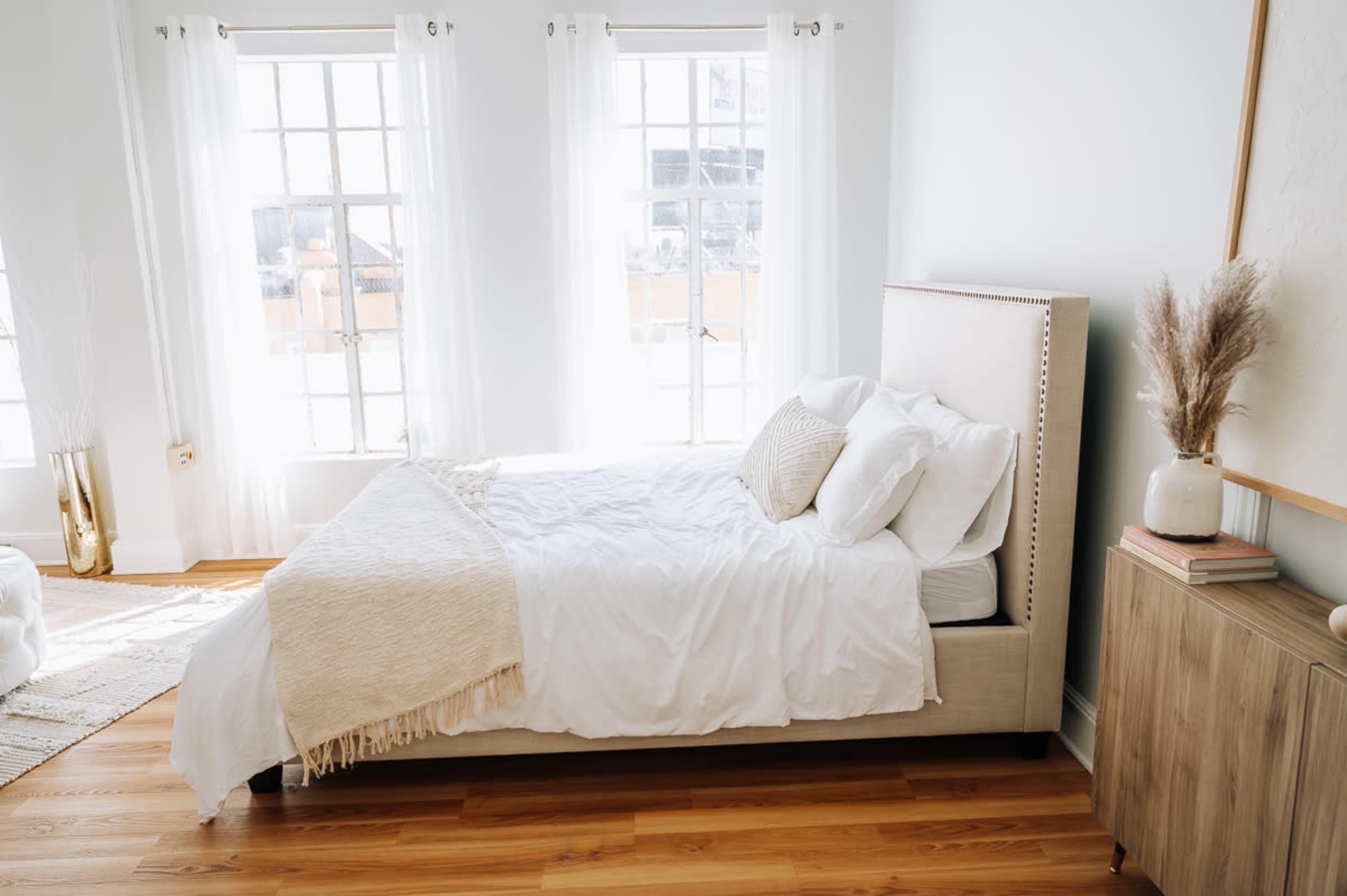 The image shows a bright bedroom featuring a bed with white linens, a decorative pillow, and a light-colored nightstand beside it, all set against large windows with sheer curtains.
