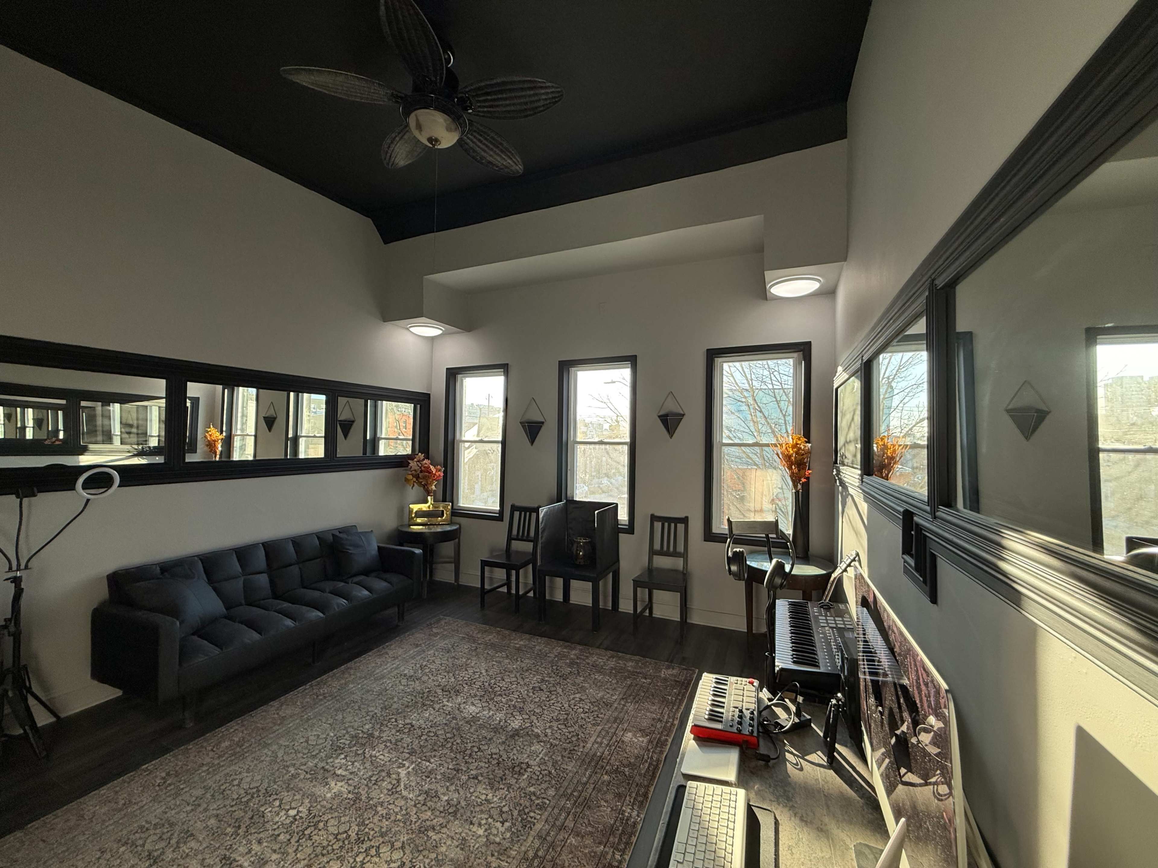The image shows a modern interior room featuring a black sofa, two black chairs, a keyboard on a desk, and large windows with natural light illuminating the space.