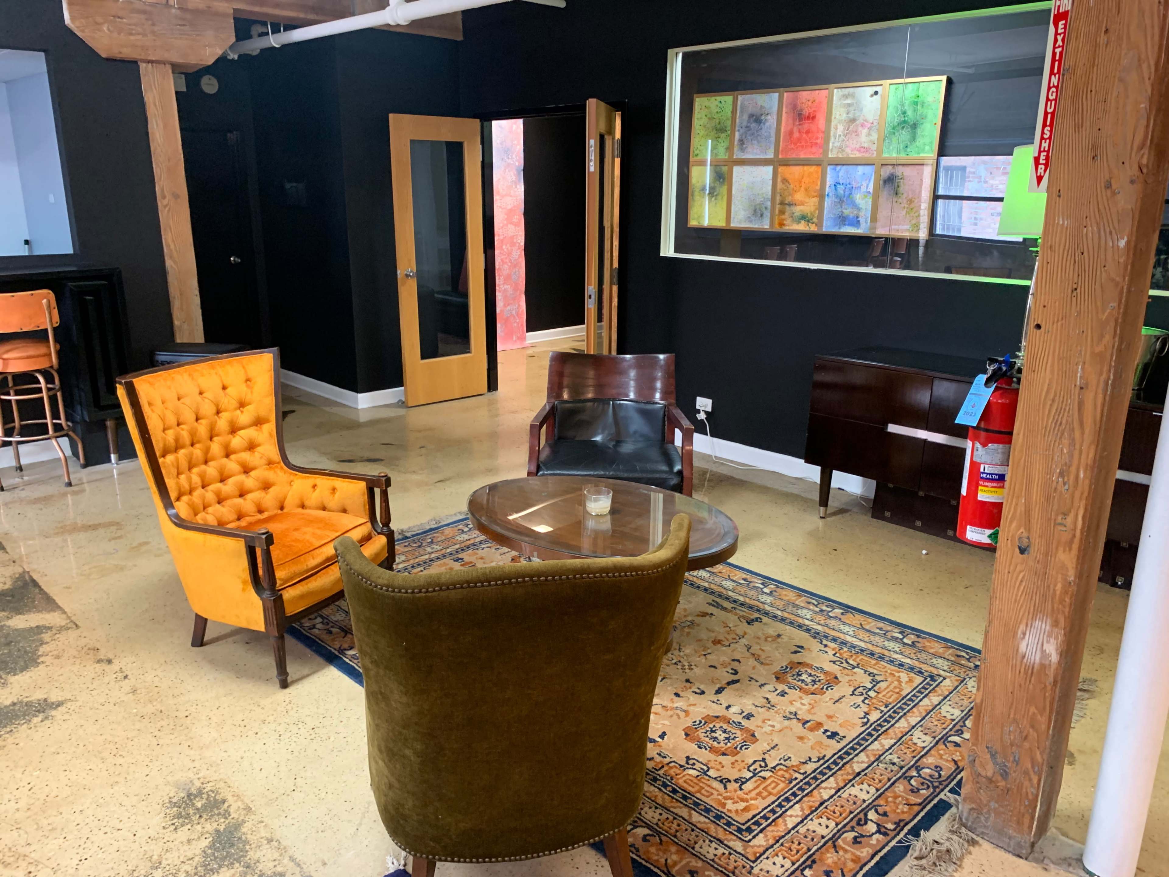 A modern interior space featuring a small seating area with two armchairs, a glass coffee table, an area rug, and a wall displaying colorful art pieces.