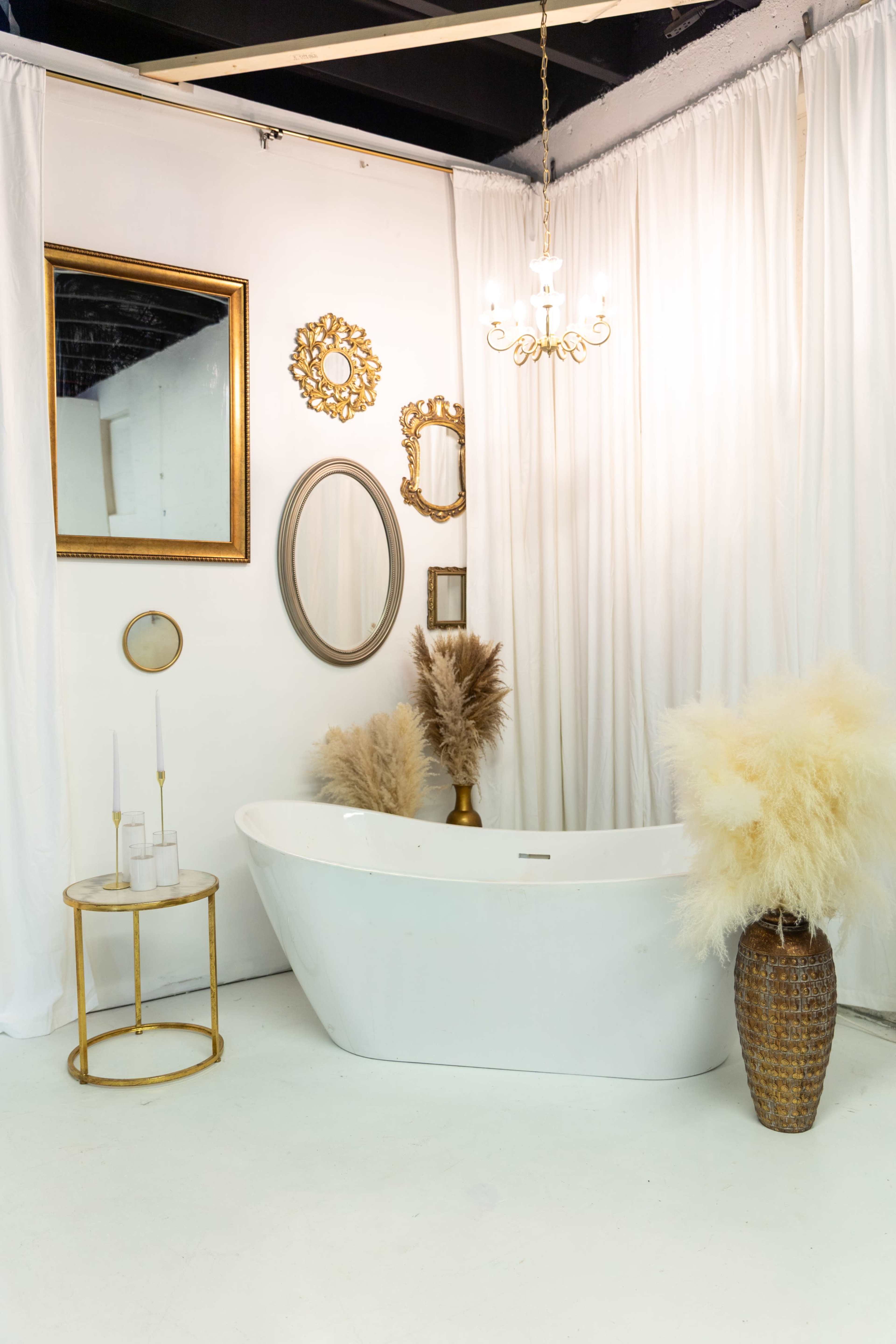 The image shows a stylish bathroom setting featuring a freestanding white bathtub, decorated with various framed mirrors and golden accents, alongside a table with candles and a large vase containing decorative pampas grass.