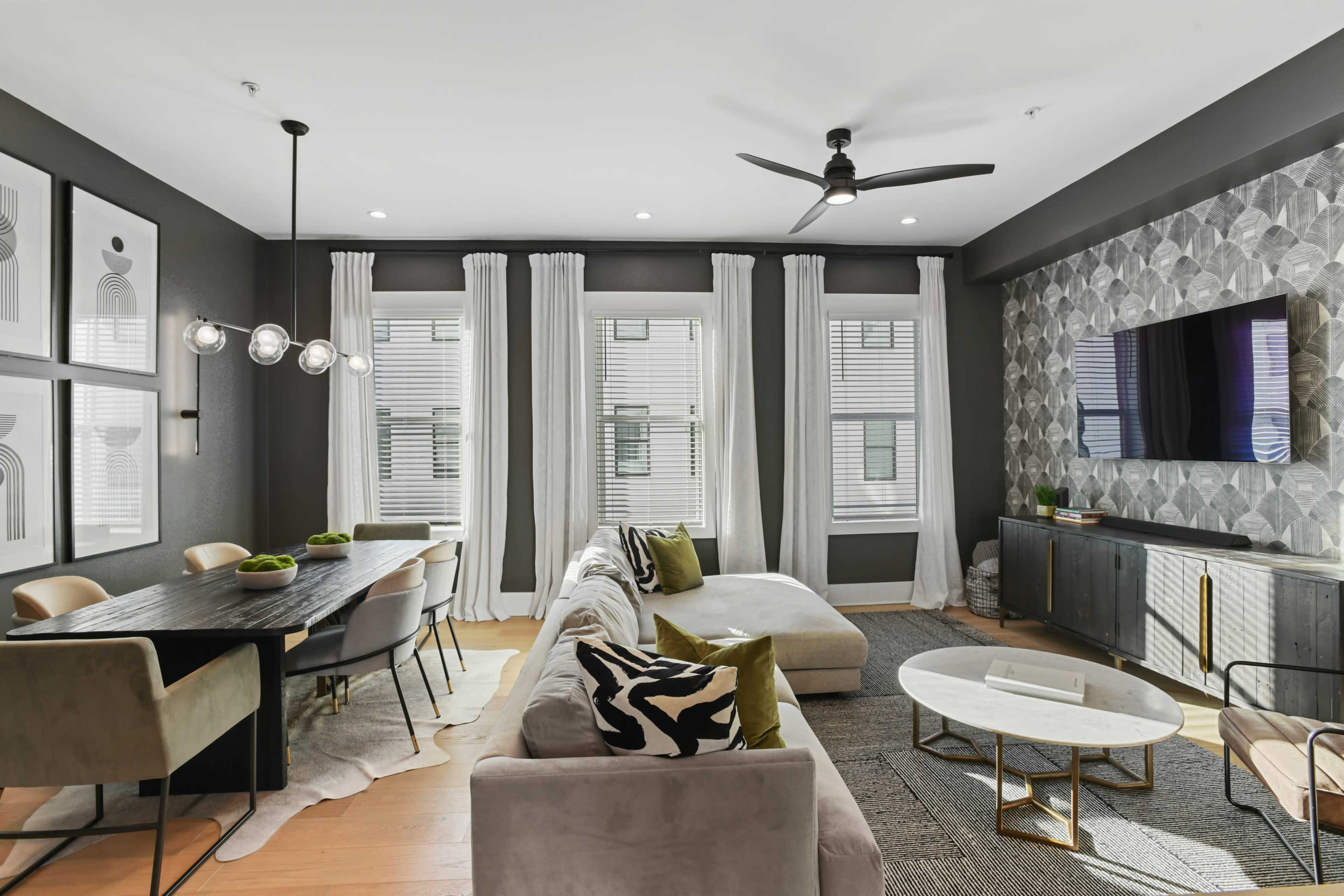 The living area features a modern design with a sectional sofa, a dining table, and a wall-mounted TV, surrounded by large windows and decorative wall art.