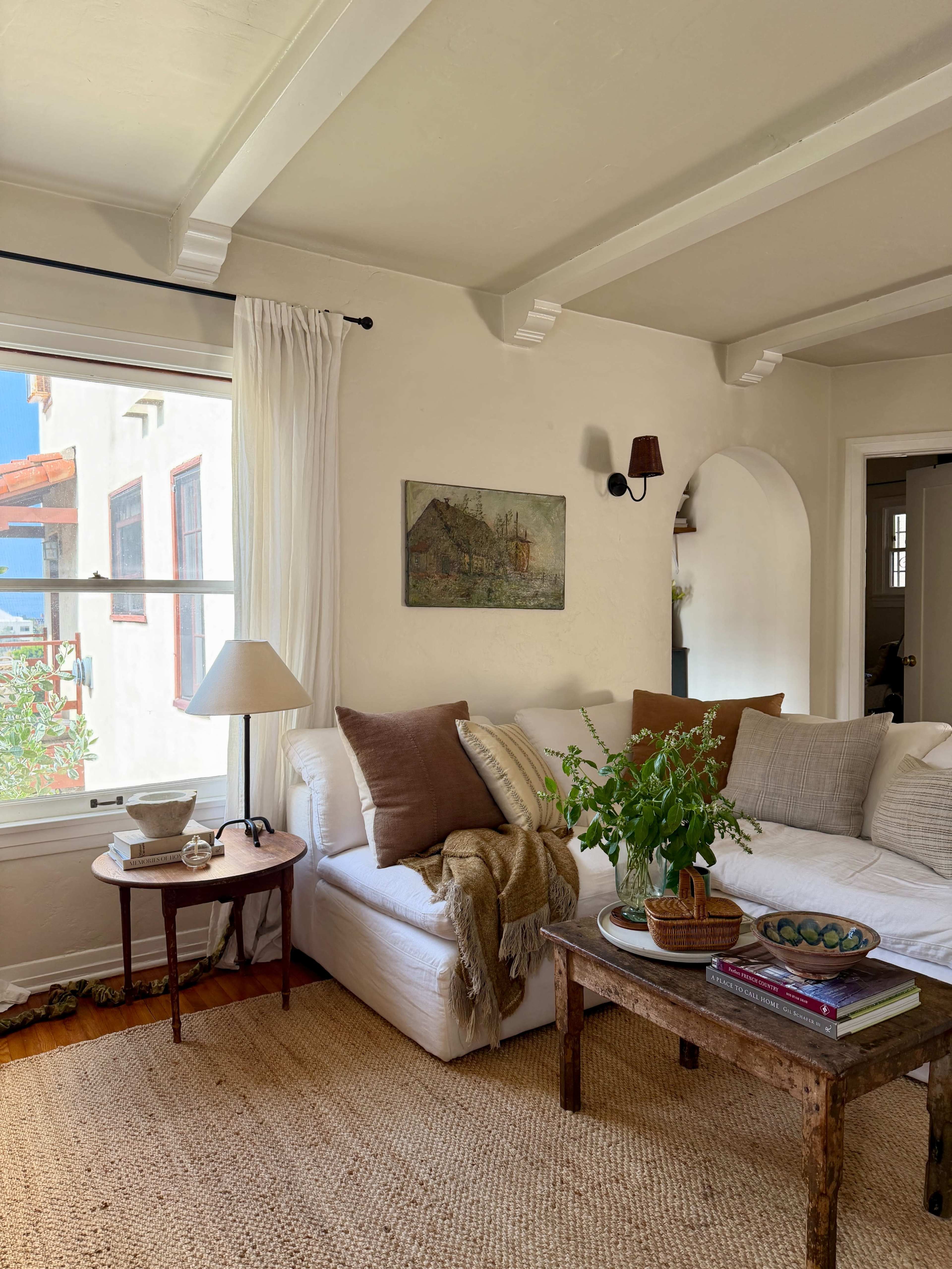 The image shows a cozy living room featuring a white sofa adorned with brown cushions, a wooden coffee table, a floor lamp, and a large window letting in natural light.