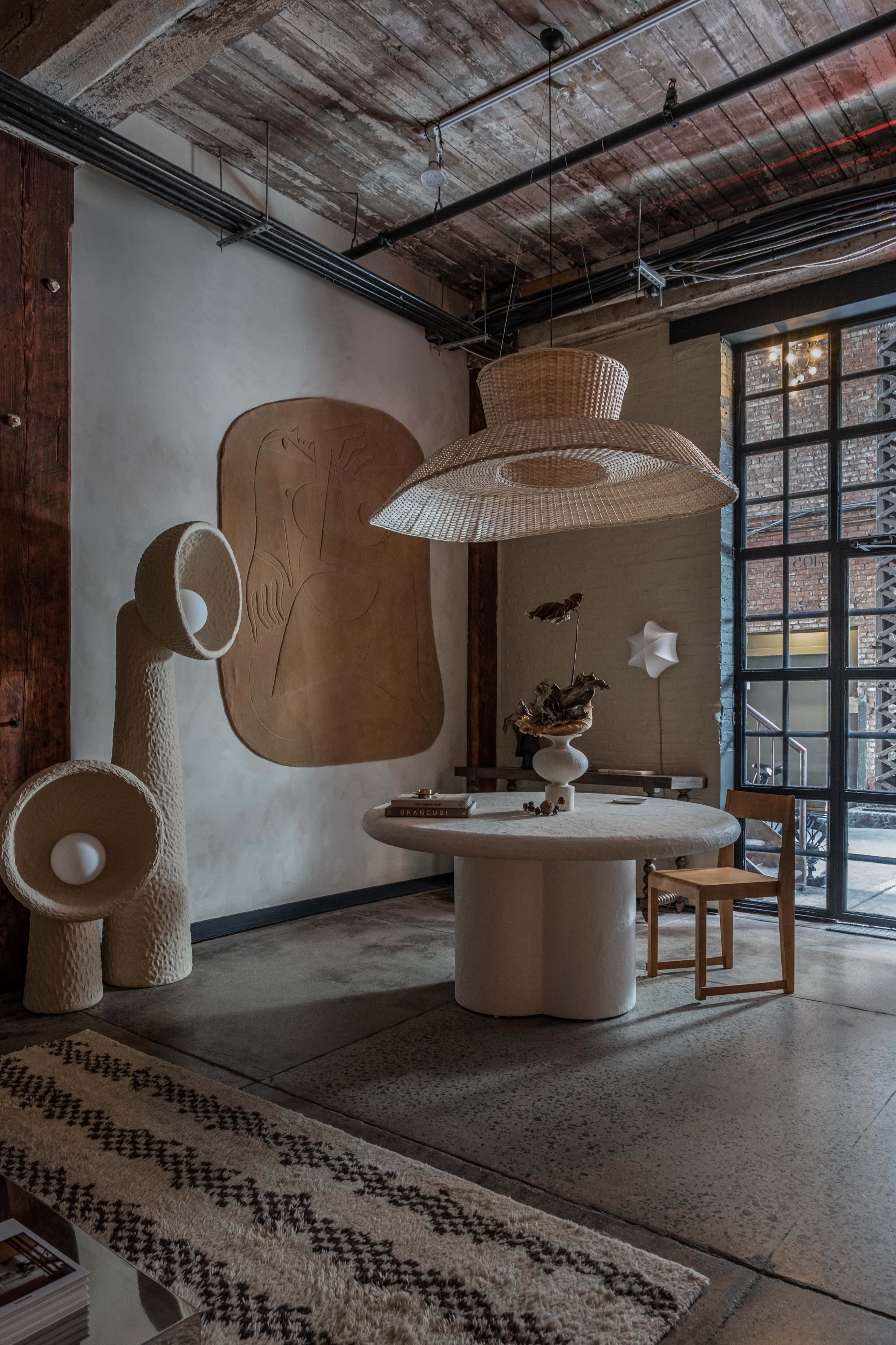A stylish interior featuring a circular table, a woven hanging light fixture, and sculptural decor against a textured wall.
