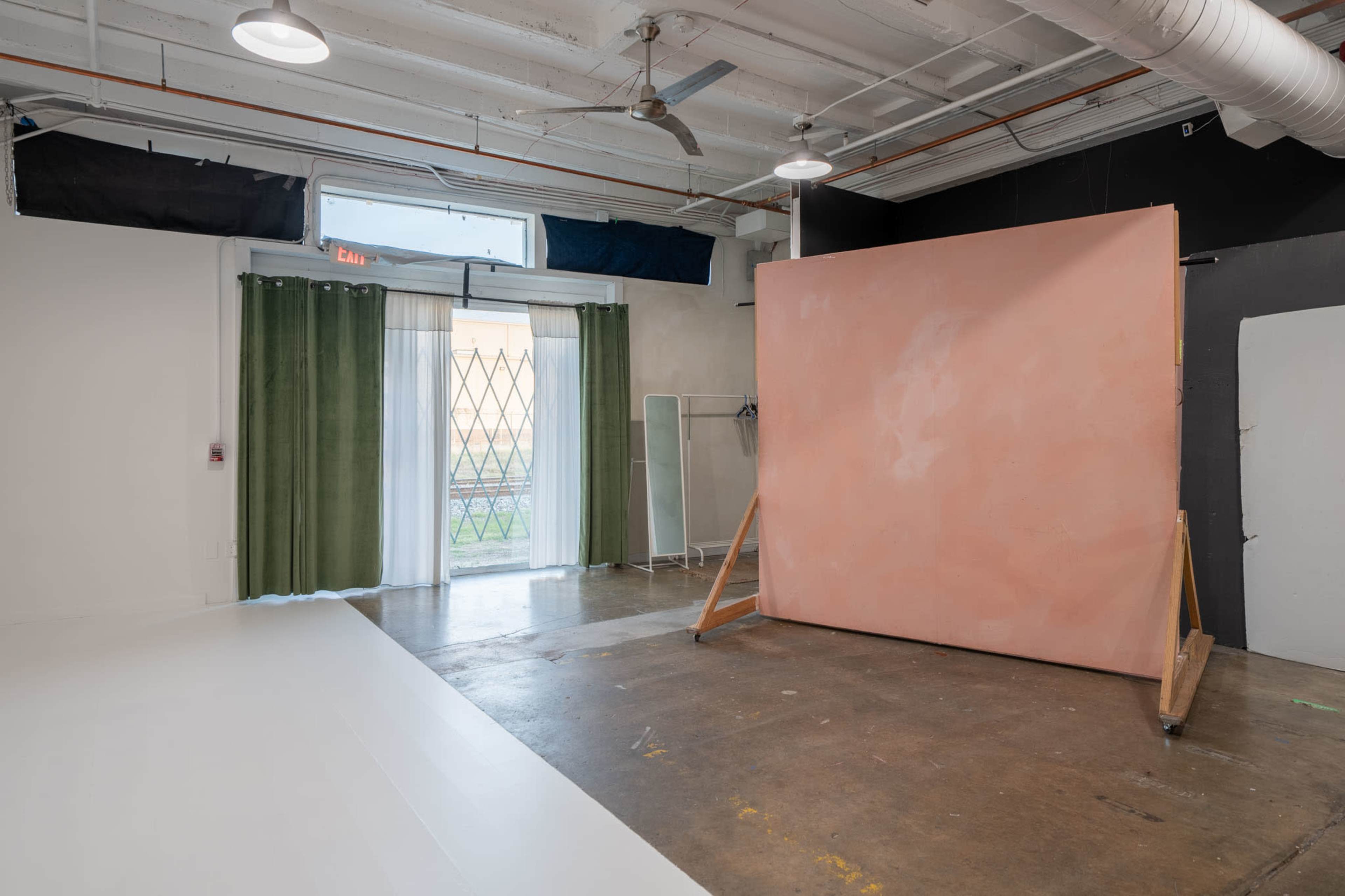 The image shows a spacious studio with a large pink backdrop, a window with green curtains, and concrete flooring.