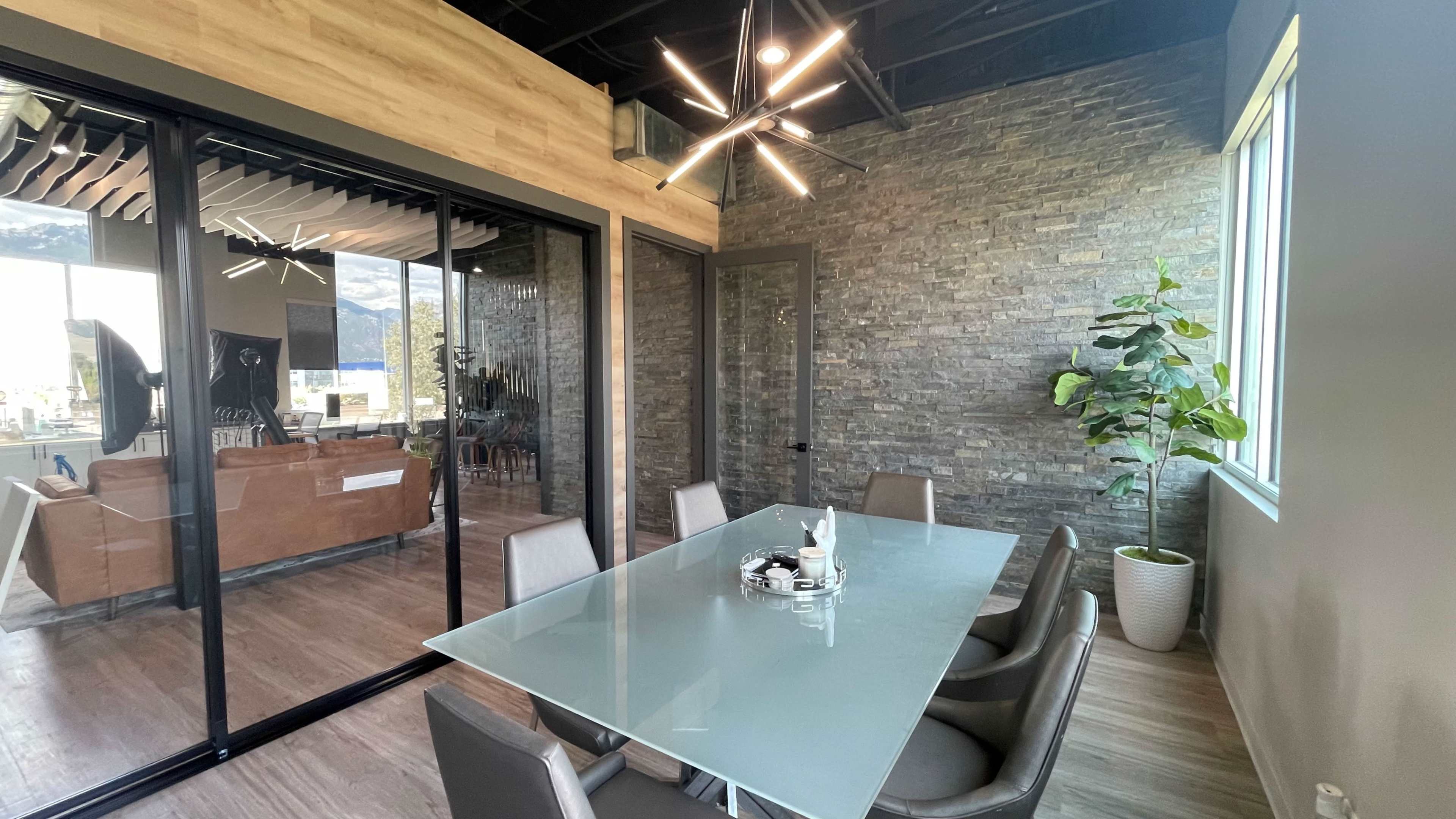 A modern conference room features a glass table surrounded by chairs, with a stone accent wall and a large window overlooking an adjacent space.