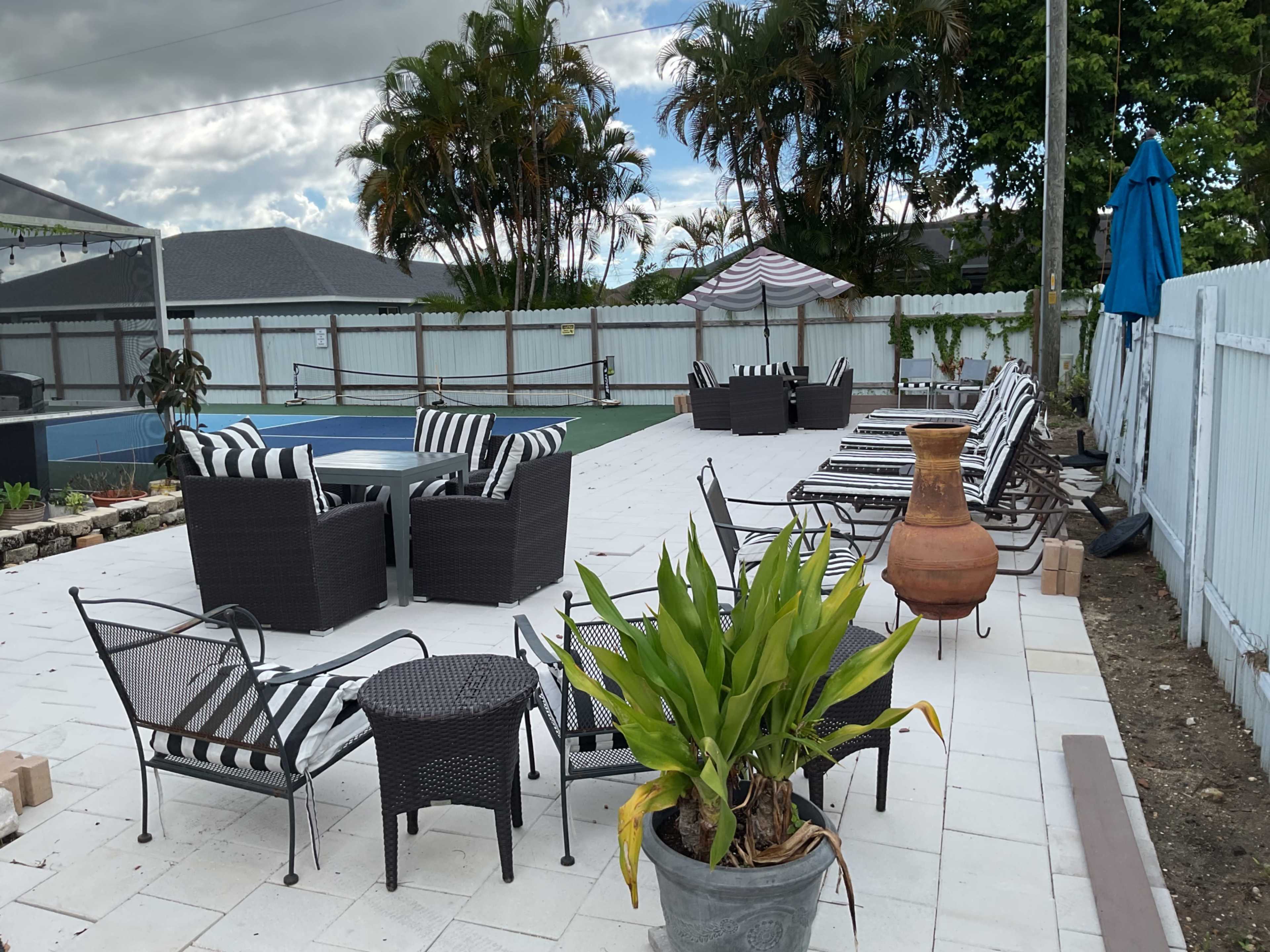 The image depicts a poolside area with lounge chairs arranged around a table, surrounded by palm trees and outdoor decor.