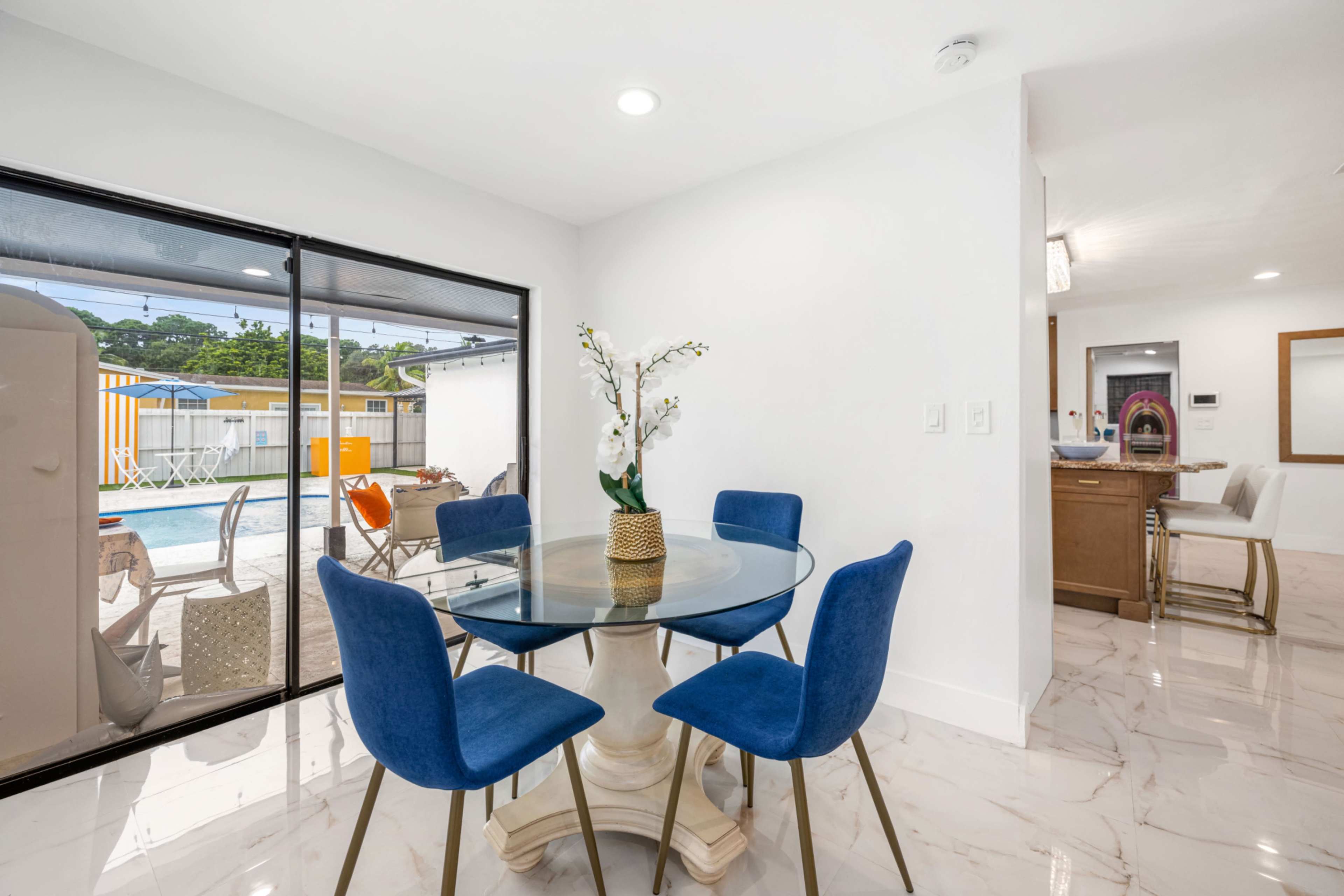 A glass dining table surrounded by blue chairs is situated near large windows that overlook a pool area.