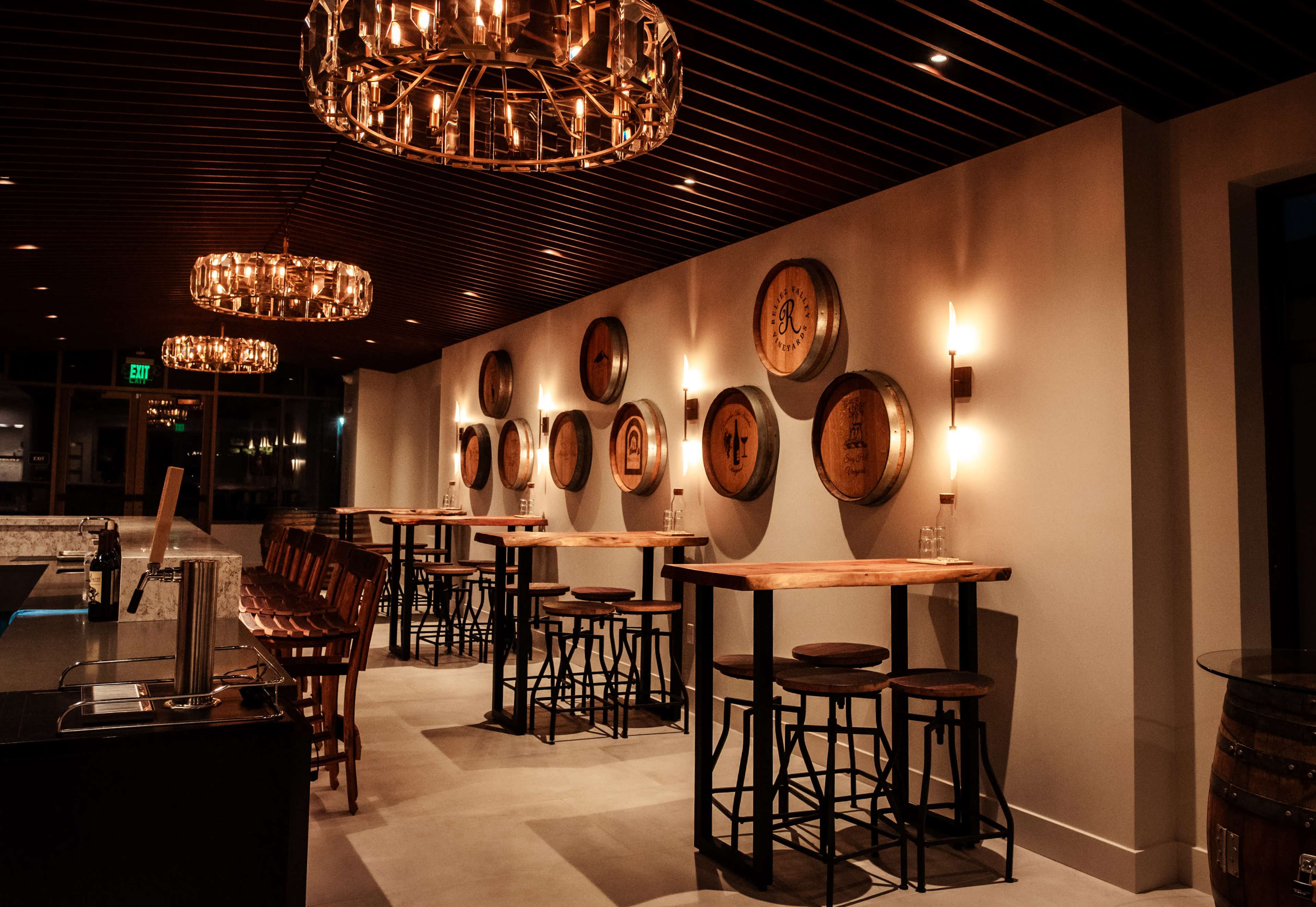 The image shows a modern bar interior with wooden tables, high stools, and decorative wall-mounted barrels.