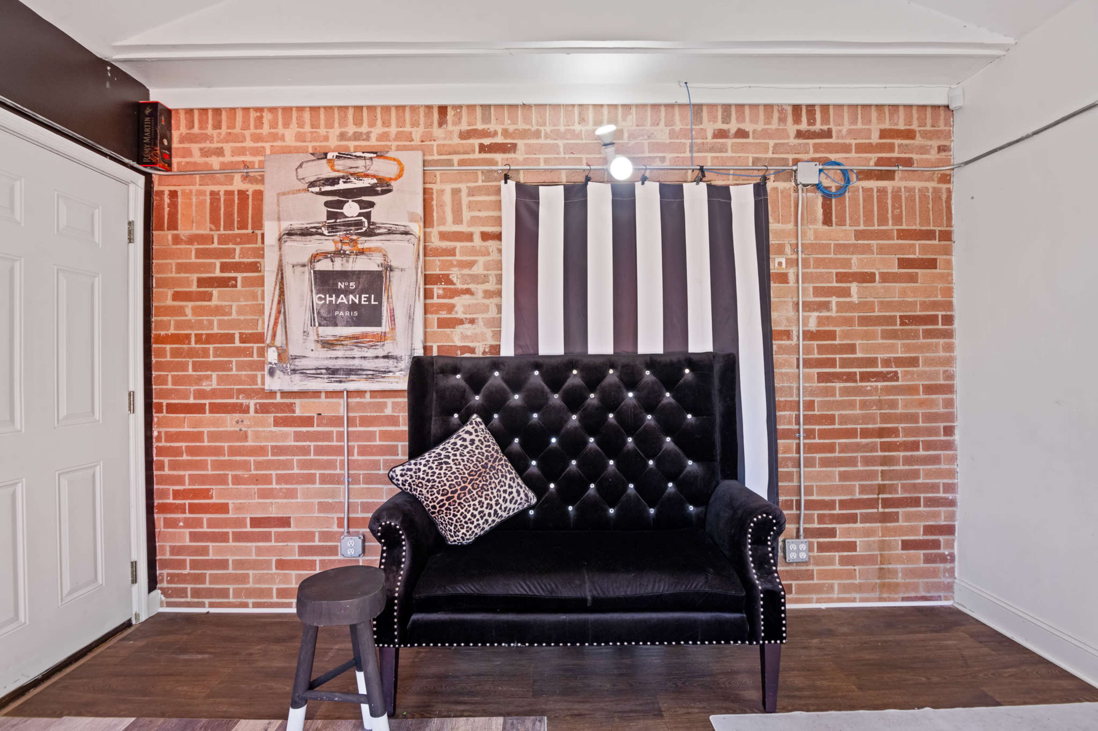 The image shows a black upholstered couch with a decorative pillow in front of a brick wall, accompanied by a striped curtain and an art piece featuring a Chanel-themed design.