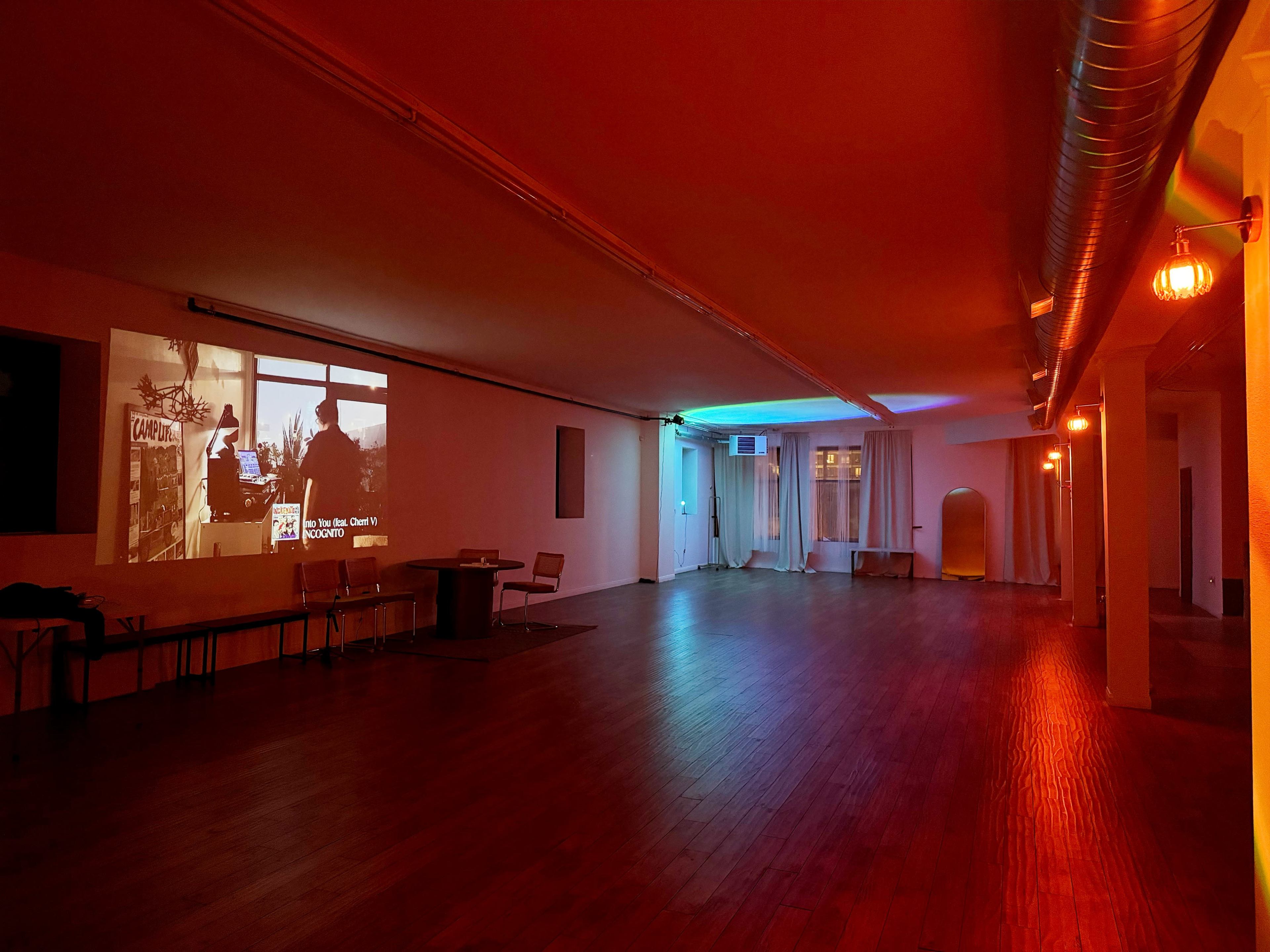 An empty room with wooden flooring, ambient colored lighting, and a projection on the wall showing a scene from a movie.