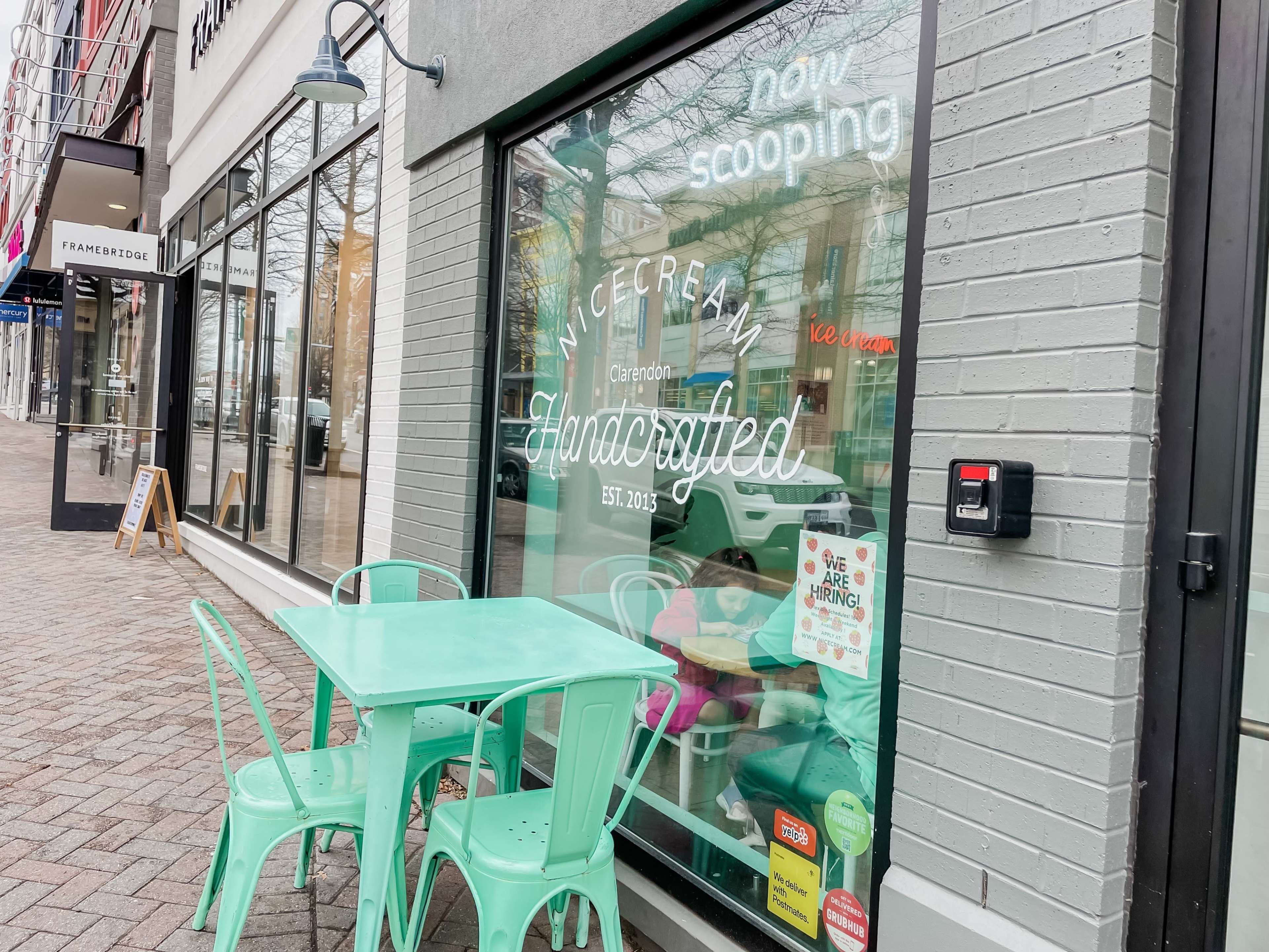 The image shows an ice cream shop with a large window display that includes the text "now scooping" and "Handcrafted" in a modern font, along with a small table and chairs outside.