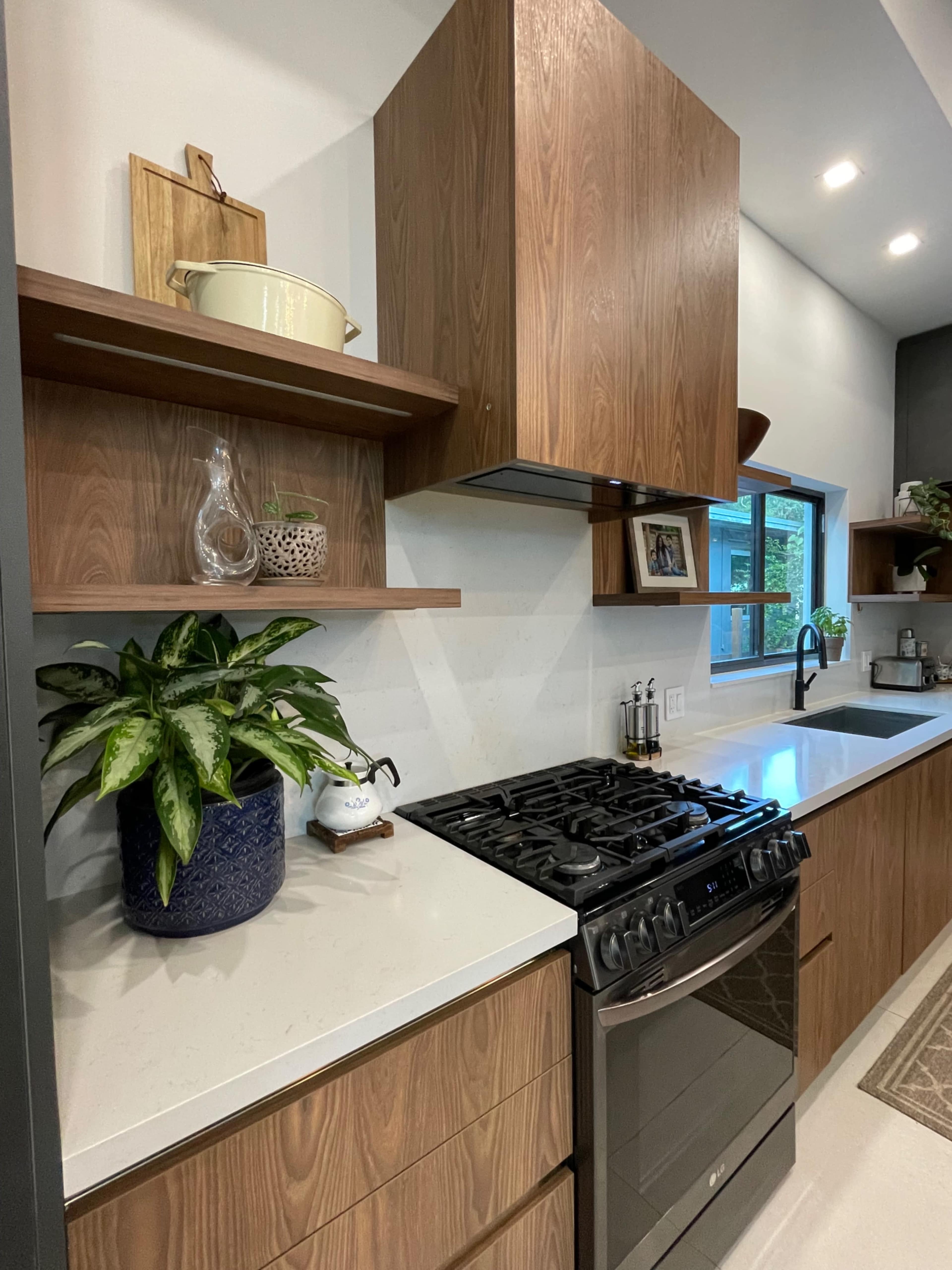 The image shows a modern kitchen featuring wooden cabinetry, a gas stove, and a window above the sink with a potted plant nearby.