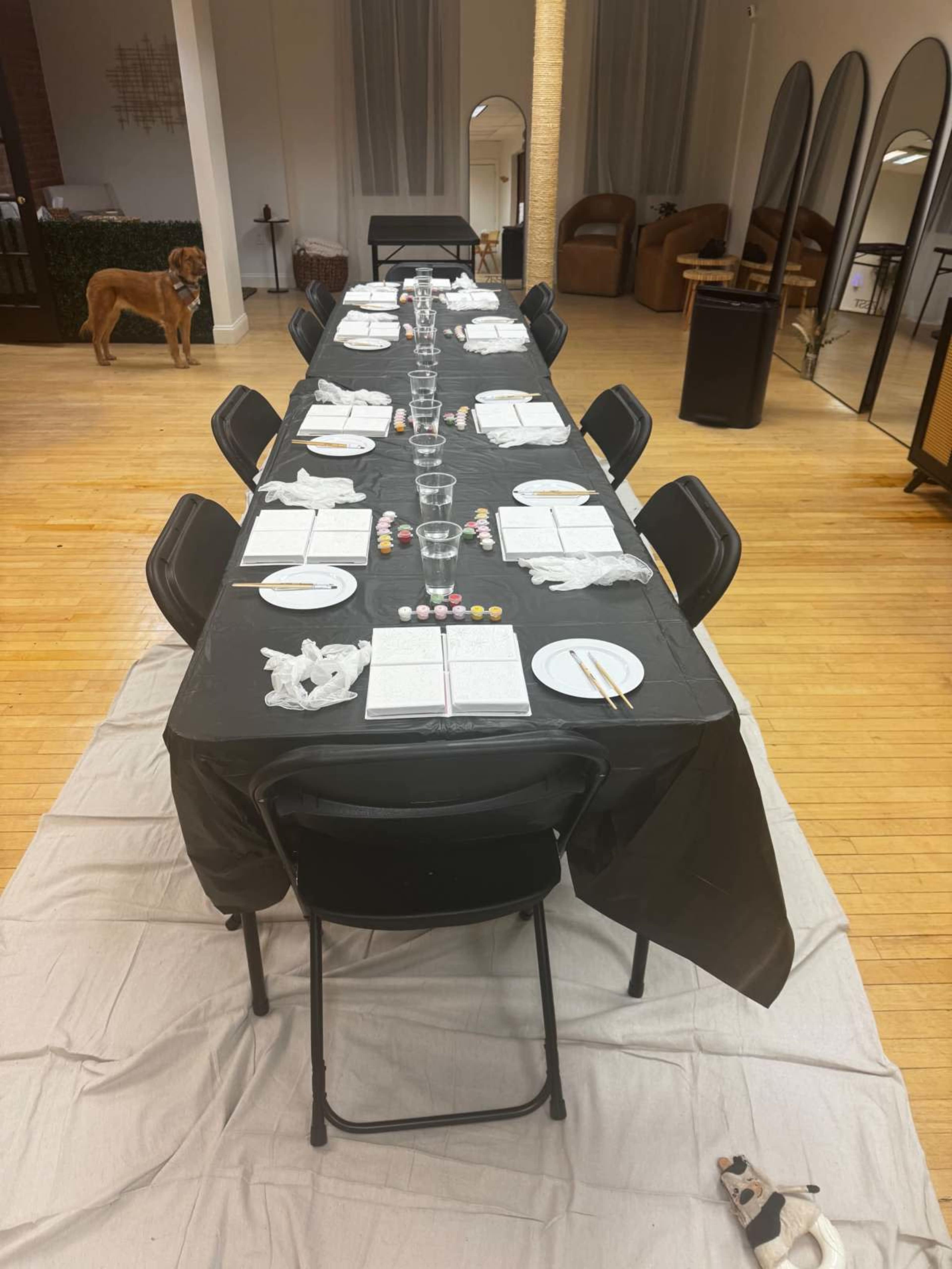 A long table is set with black tablecloths and white plates, surrounded by chairs, in a spacious room with a dog in the background.