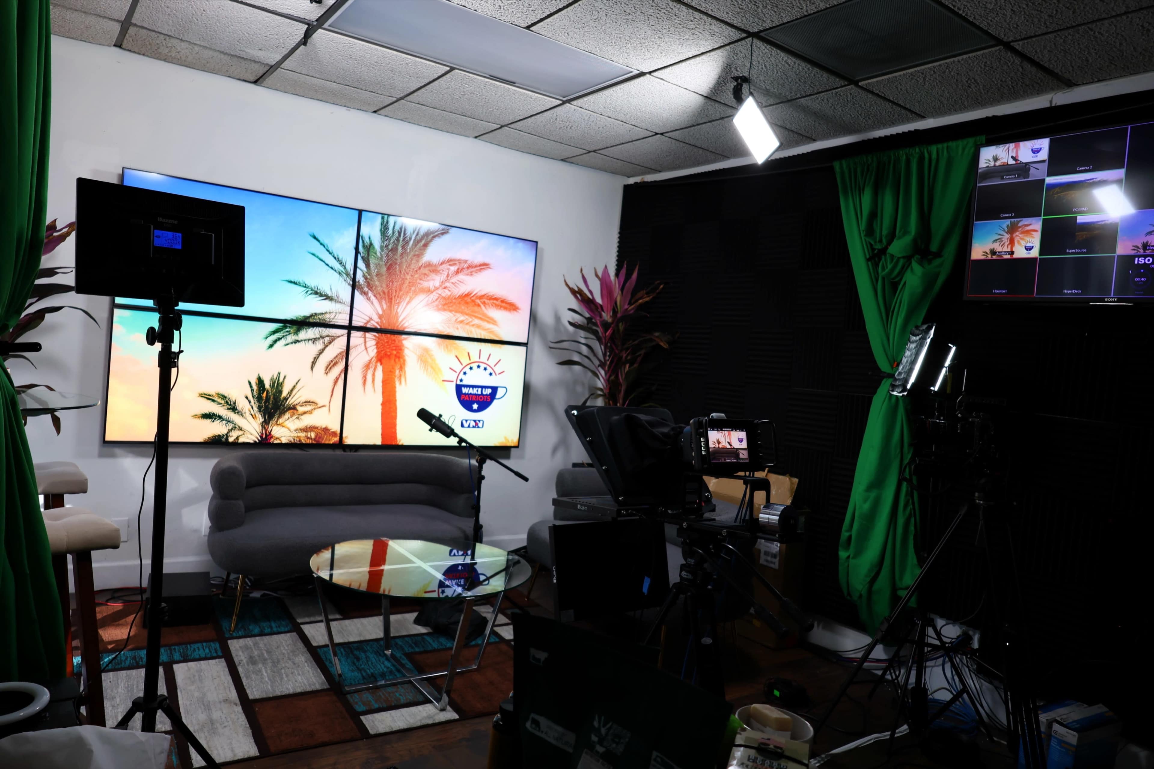 A television studio setup featuring a large screen with palm trees, a round glass table, and multiple cameras and lighting equipment.