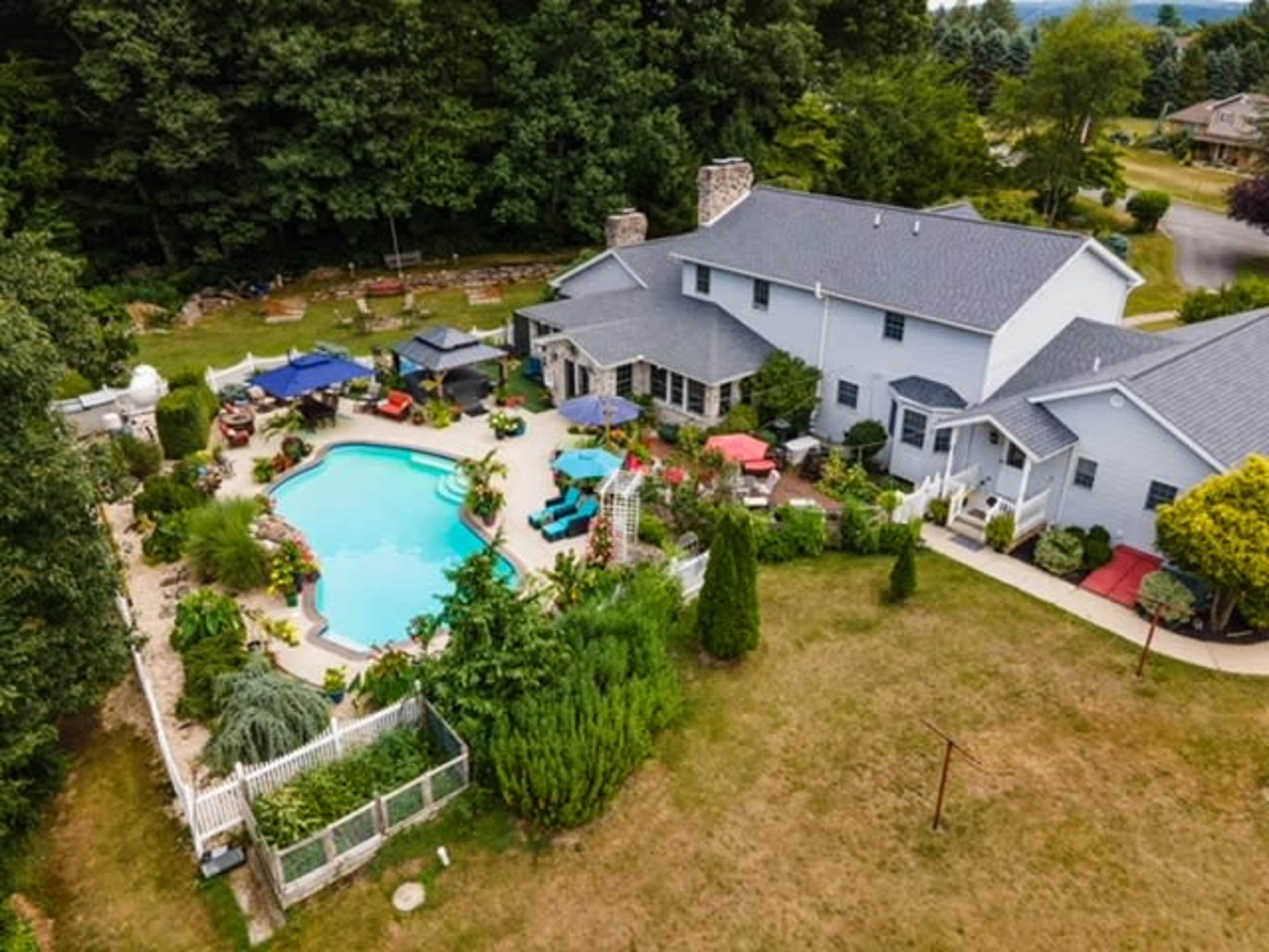 The image shows a large residential property with a swimming pool surrounded by garden features, outdoor seating, and various plants.