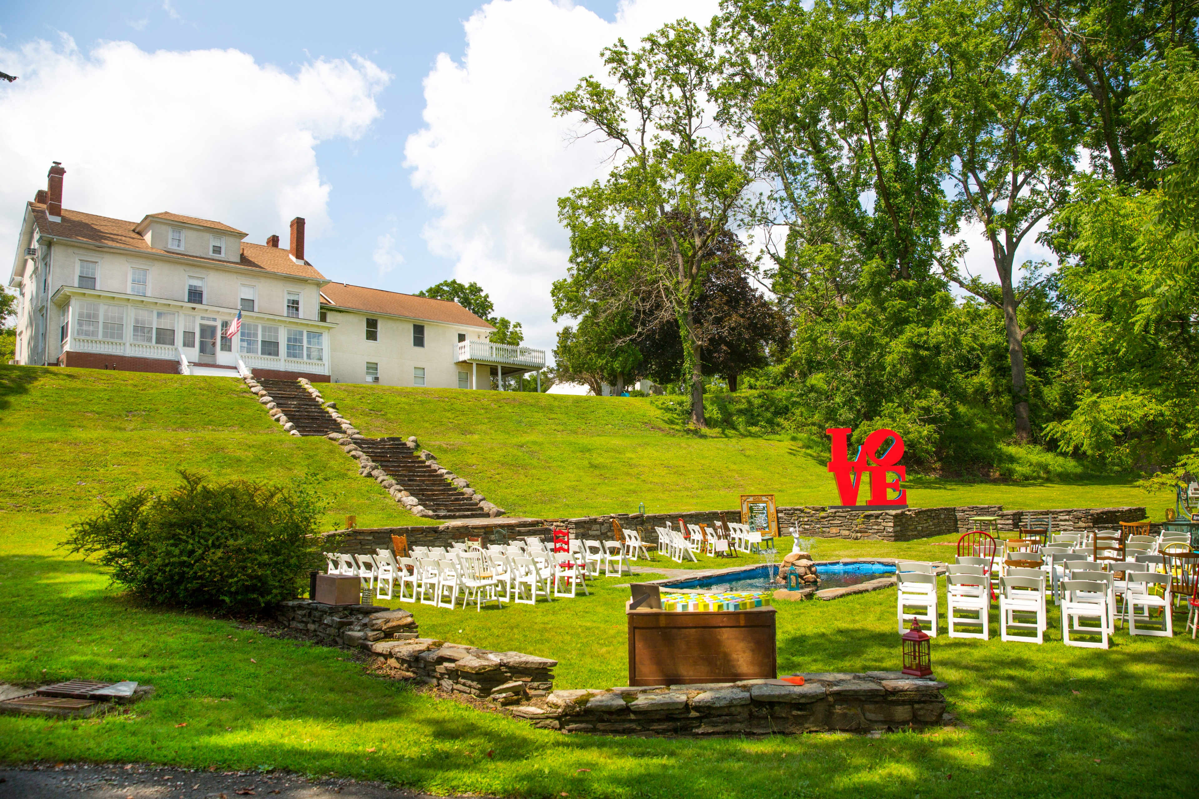 A well-maintained outdoor venue features a large green lawn with white chairs arranged in a semi-circle around a small pool and a large red "LOVE" sign in front of a multi-story white house.