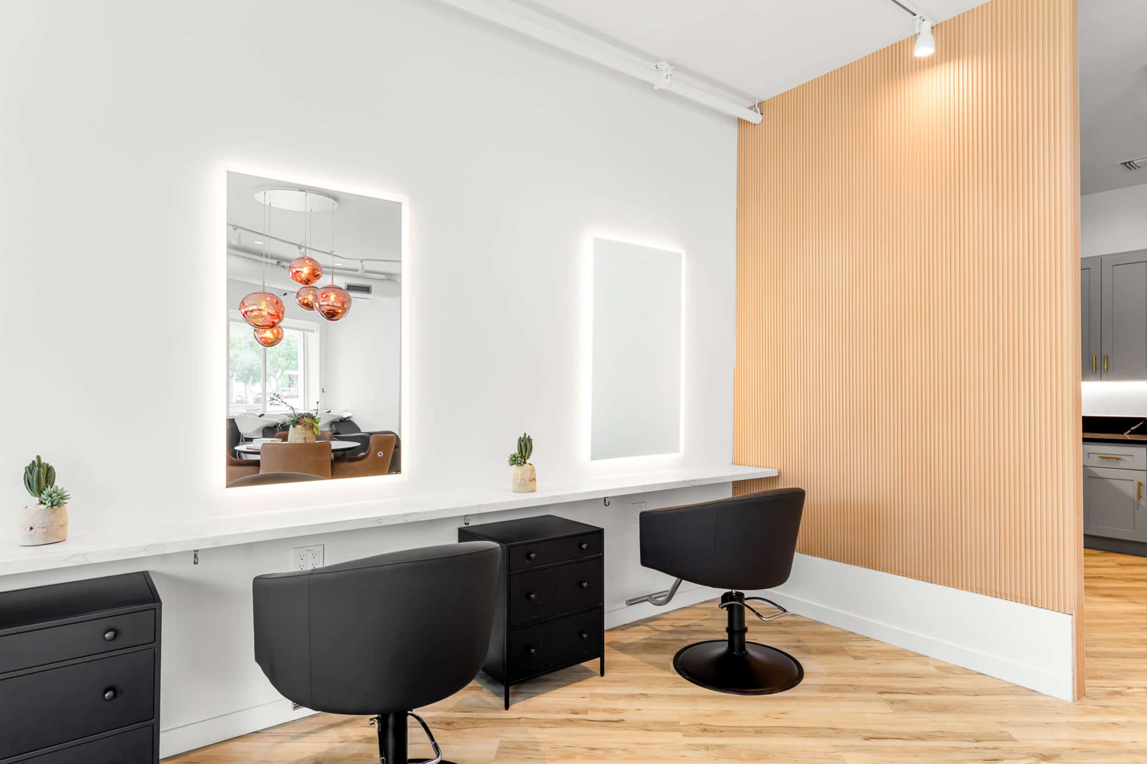 The image shows a hair salon interior with two illuminated mirrors, black salon chairs, and a beige wooden accent wall.